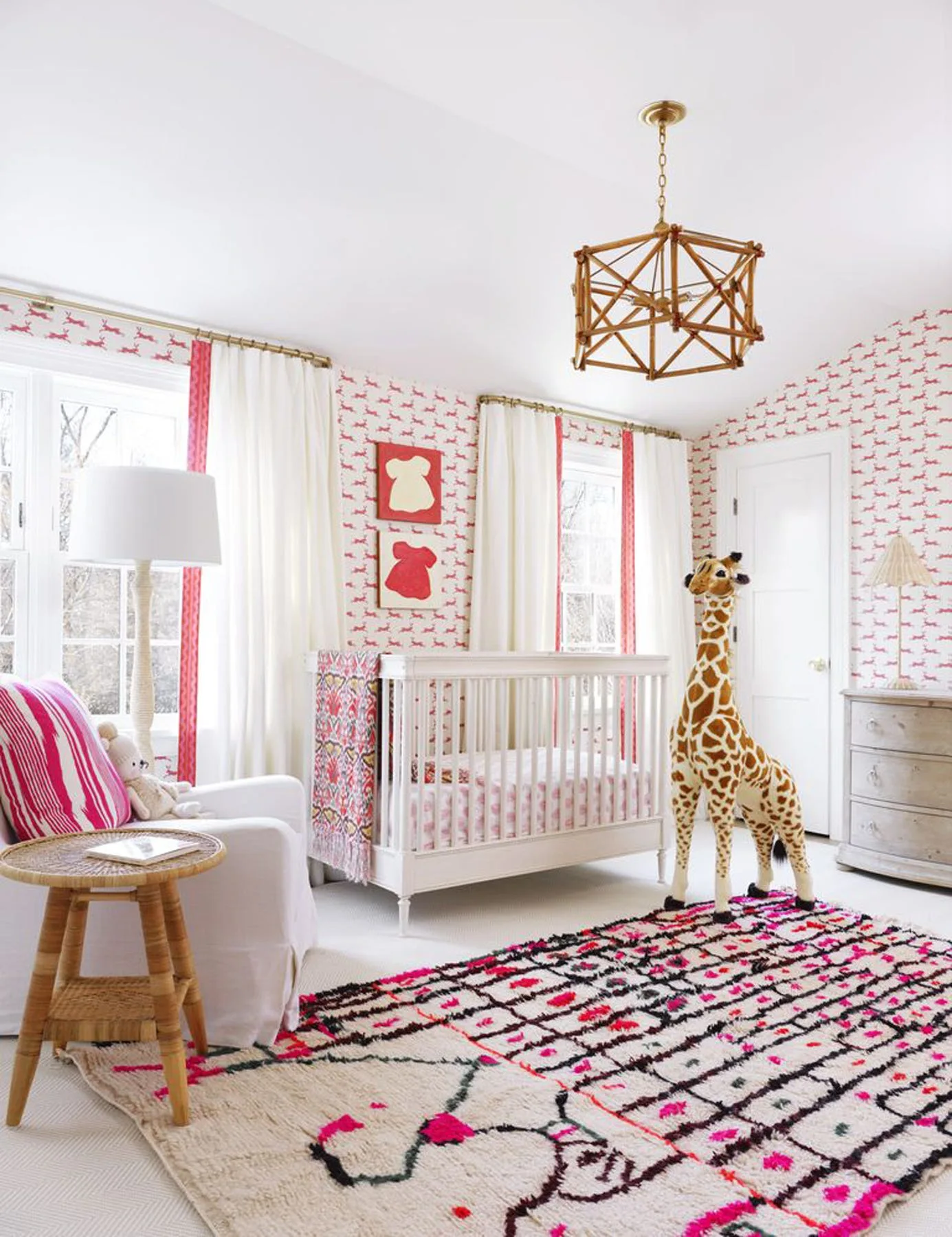 A bright nursery featuring a white crib, pink patterned wallpaper, white curtains with pink borders, a geometric rug, and a tall stuffed giraffe.