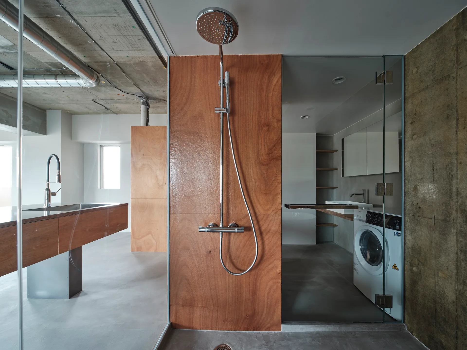An industrial bathroom features raw concrete surfaces, a warm plywood partition wall with a chrome shower, and a washer tucked under the vanity.