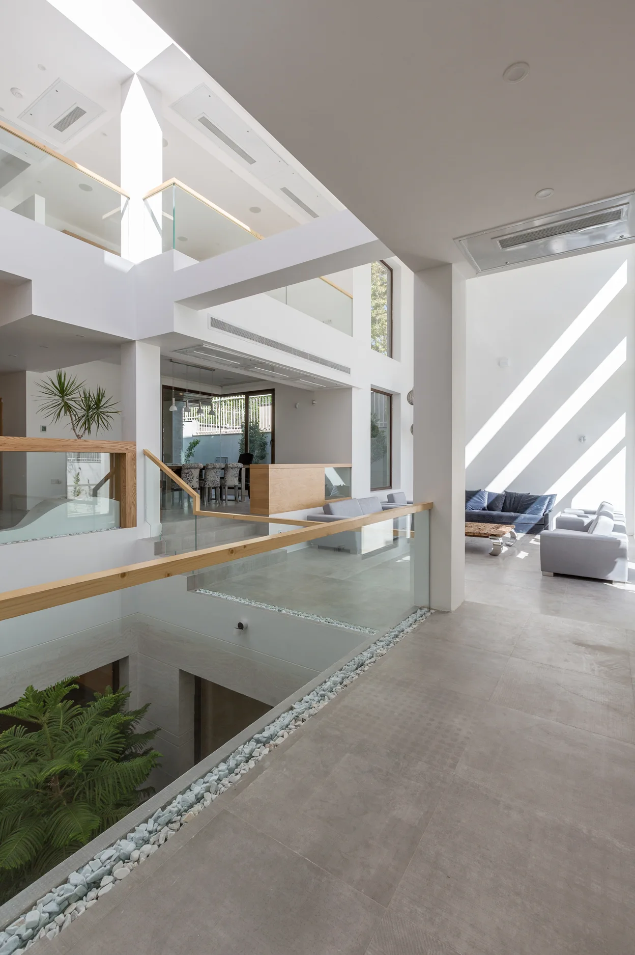 A multi-level modern interior features a central void with glass railings, light wood handrails, large grey floor tiles, and bright sunlight from a skylight.