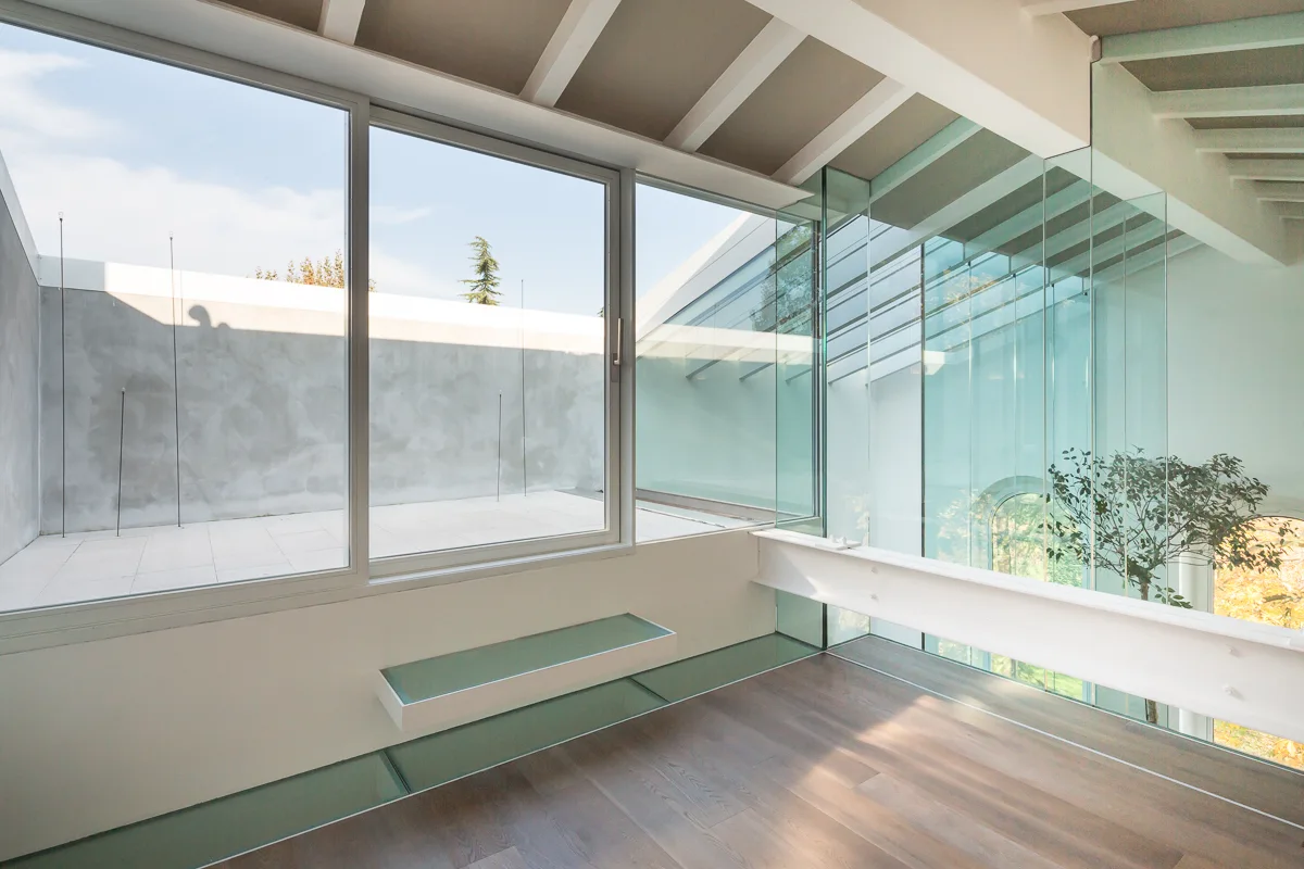 A minimalist loft space with wood floors, large glass walls showing a concrete terrace, and white exposed ceiling beams.