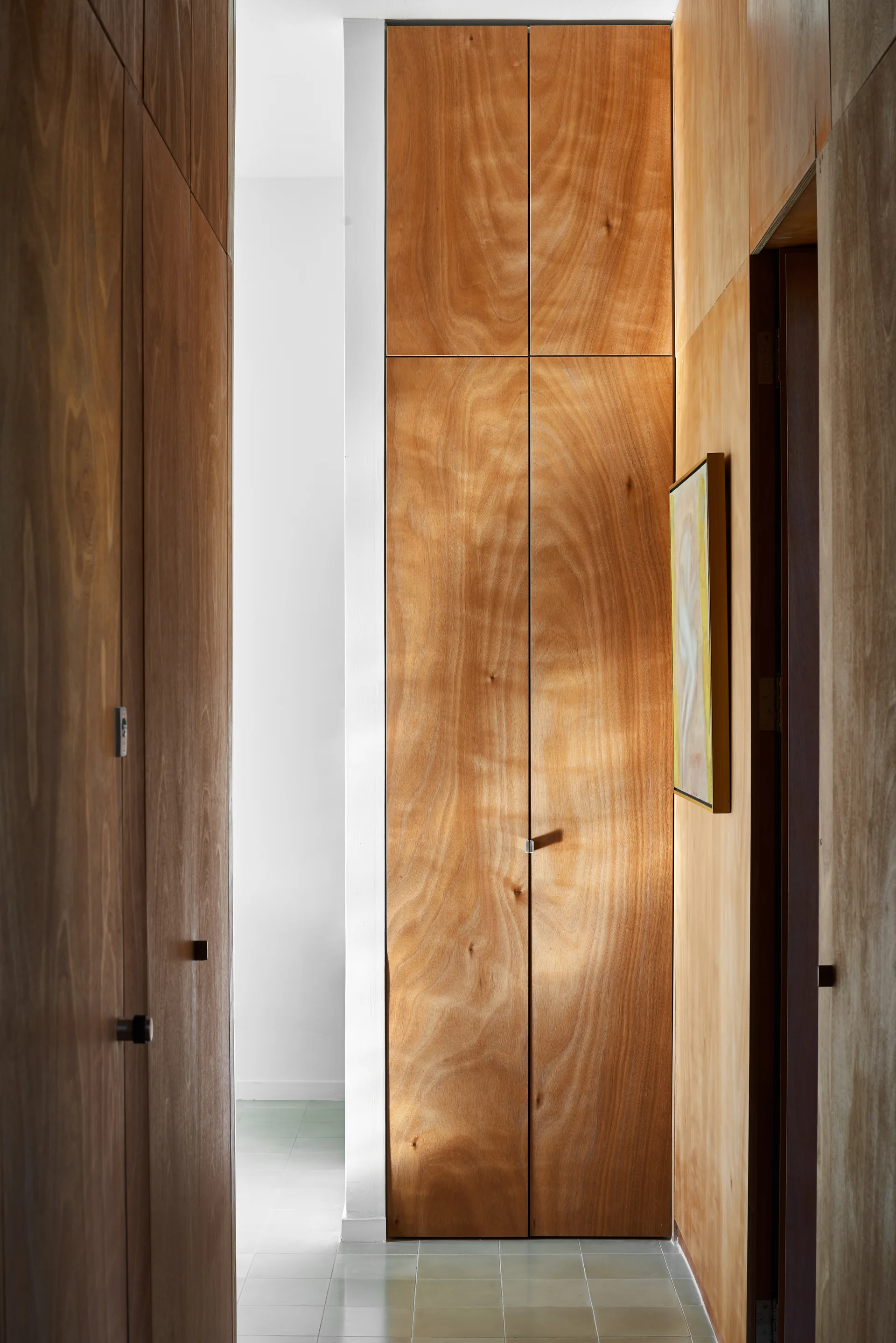 A narrow hallway lined with floor-to-ceiling warm plywood storage cabinets with clean-lined doors, leading to a bright room.