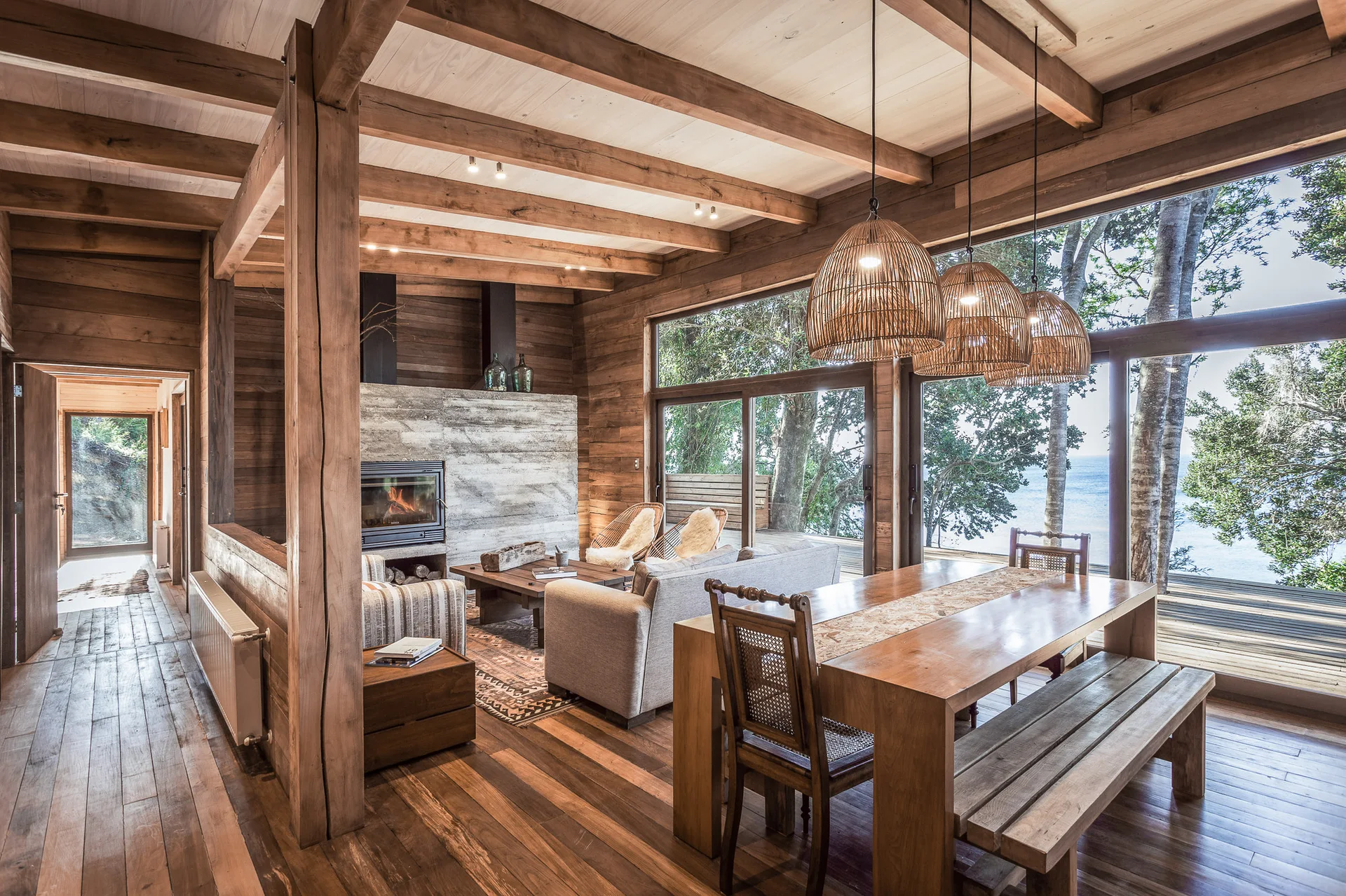 A rustic wood cabin interior features an open-plan living and dining area with exposed ceiling beams, a concrete fireplace, and large windows.