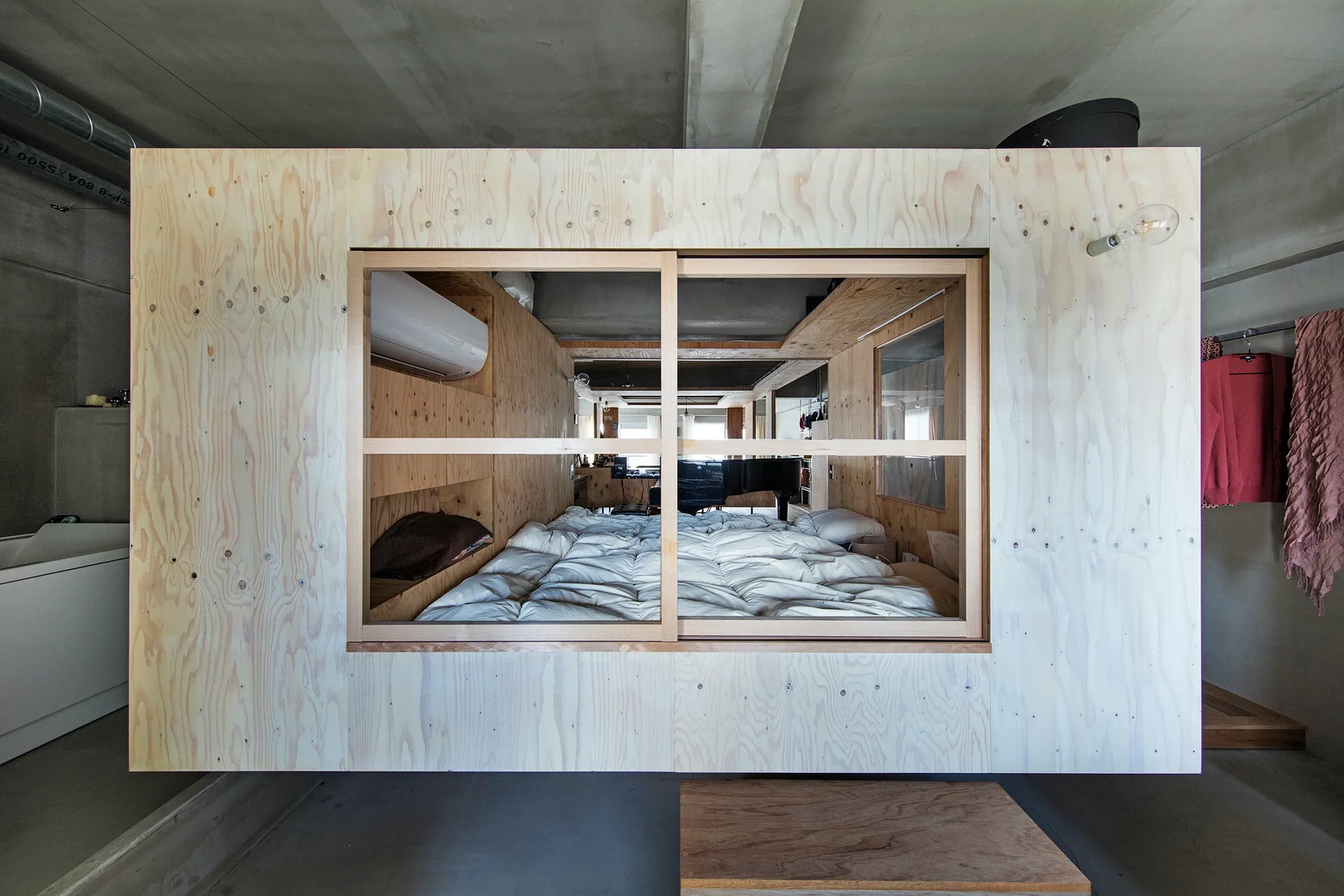A compact plywood bedroom module with a sliding glass window and white bedding sits inside an industrial loft with exposed concrete walls.