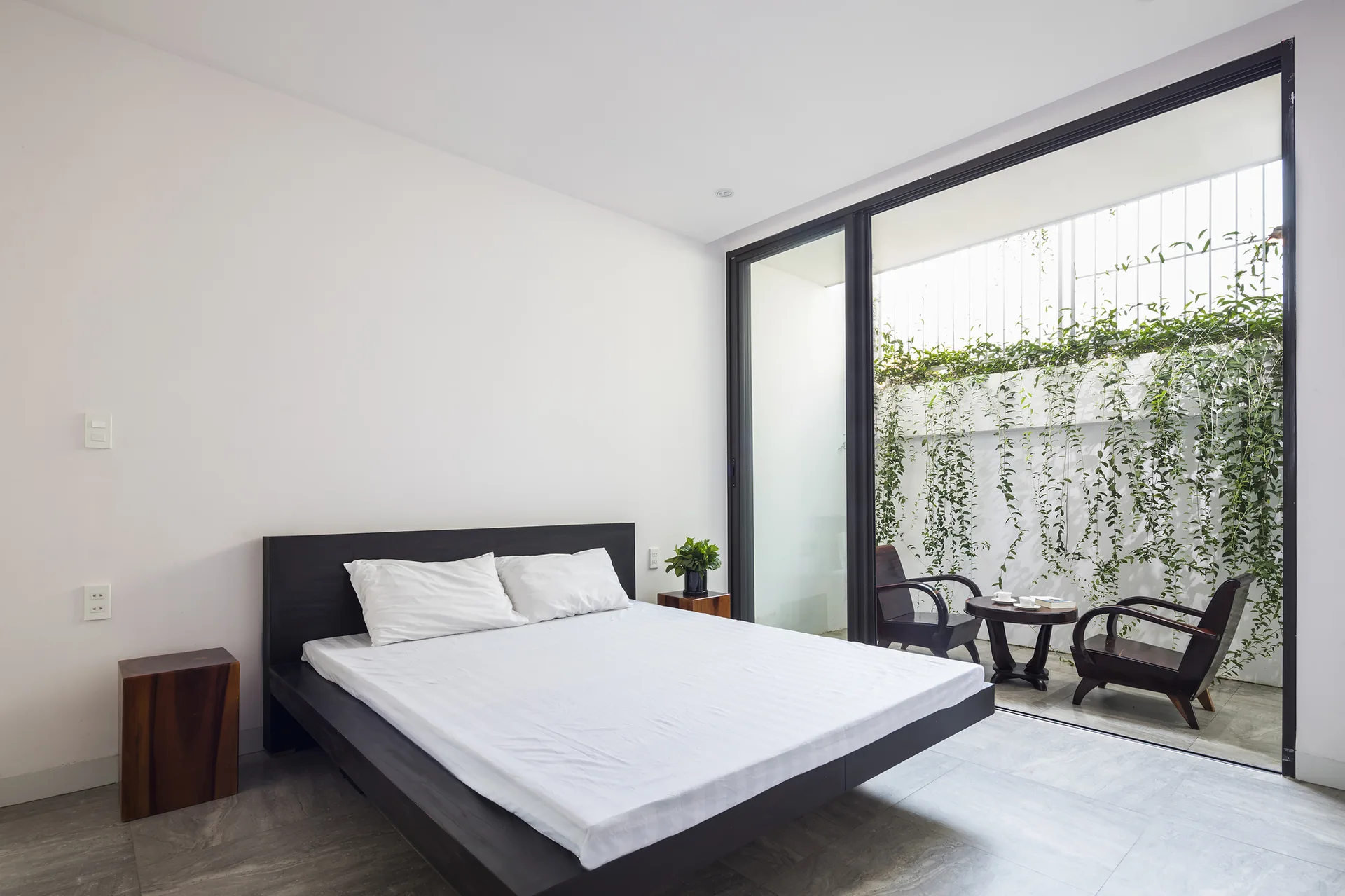 A minimalist bedroom features a dark wood platform bed with white bedding, next to a sliding glass door opening onto a patio with trailing green vines.