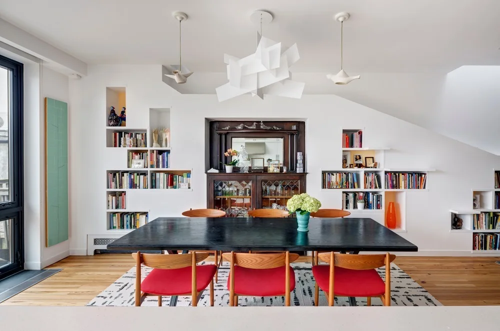 A dining room features a black wooden table with red-cushioned chairs, set before a white wall with recessed geometric bookshelves and a dark wood sideboard.