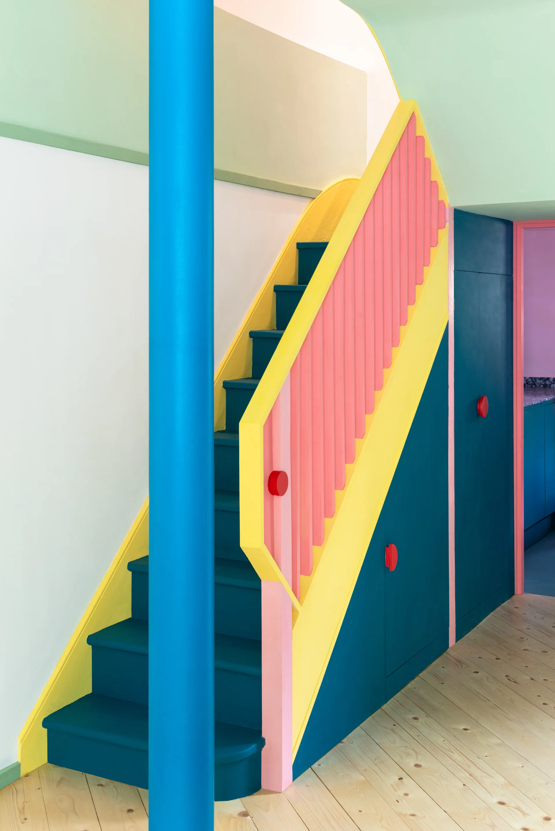 A colorful color-blocked wooden staircase featuring pink balusters, yellow handrails, teal blue steps, and a matching under-stair cabinet.
