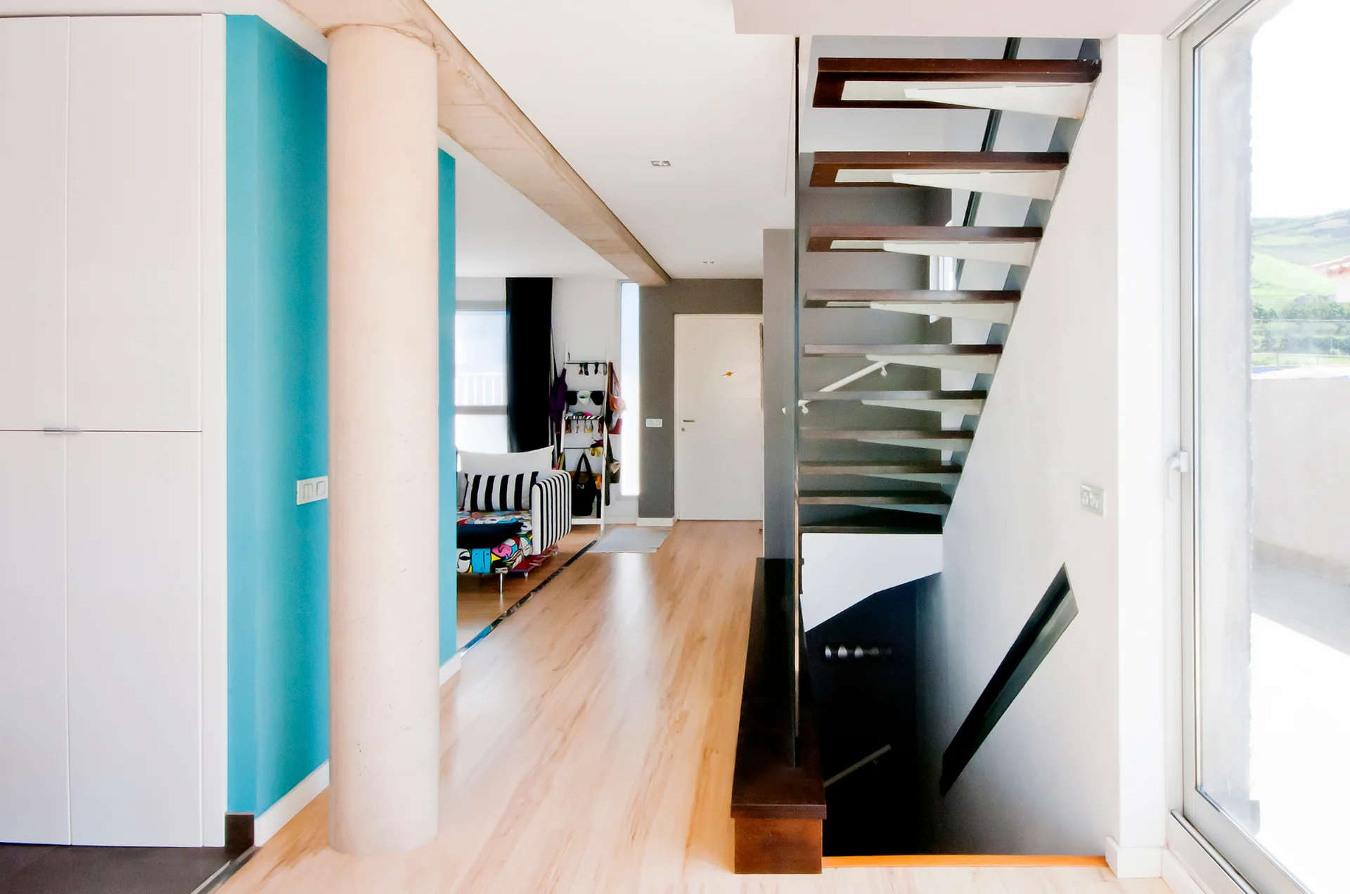A modern hallway features light wood floors, a floating staircase with dark wood treads, a round concrete column, and a bright turquoise accent wall.