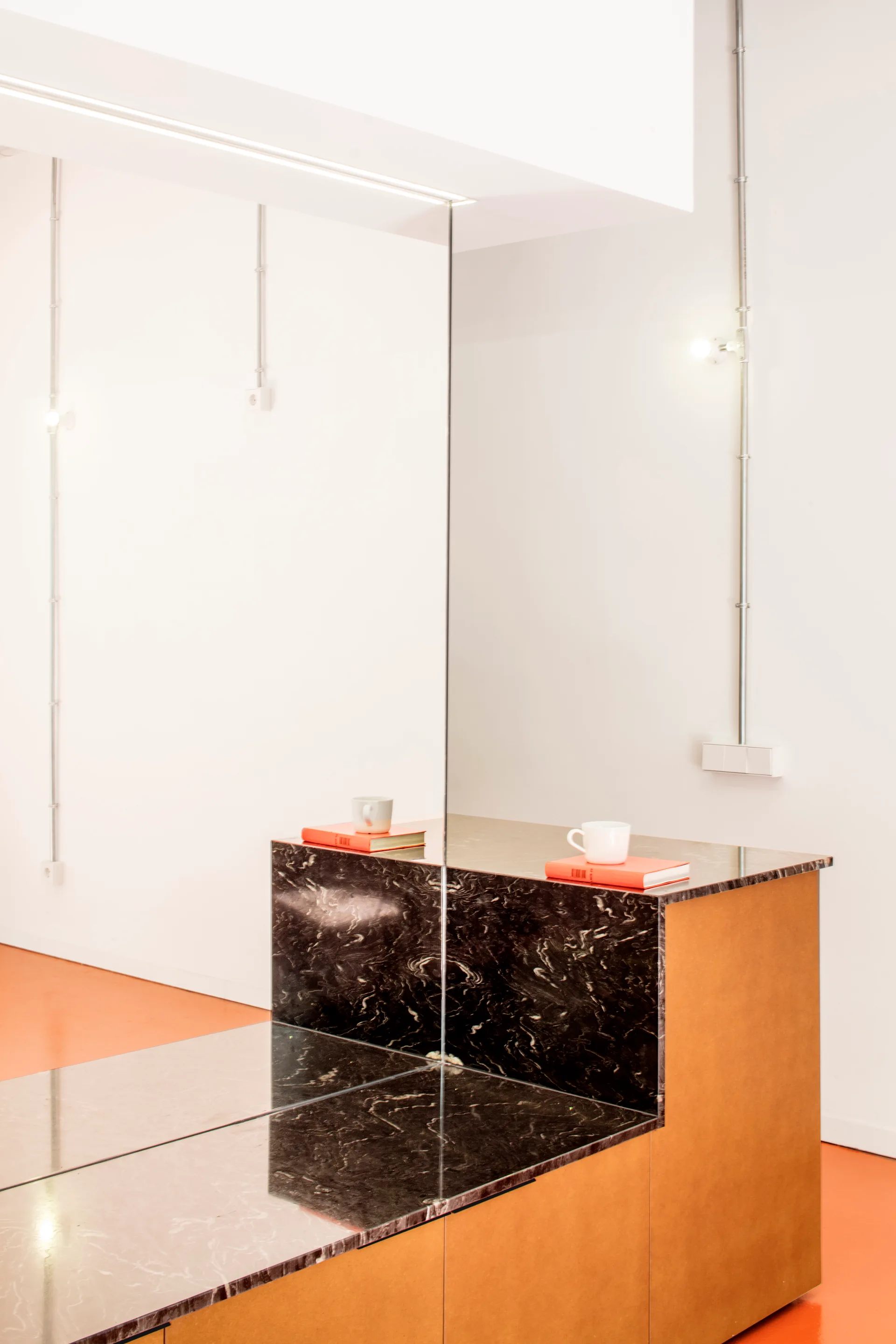 A custom cabinet with a black veined marble top sits next to a large mirror on an orange floor against white walls with exposed conduit.