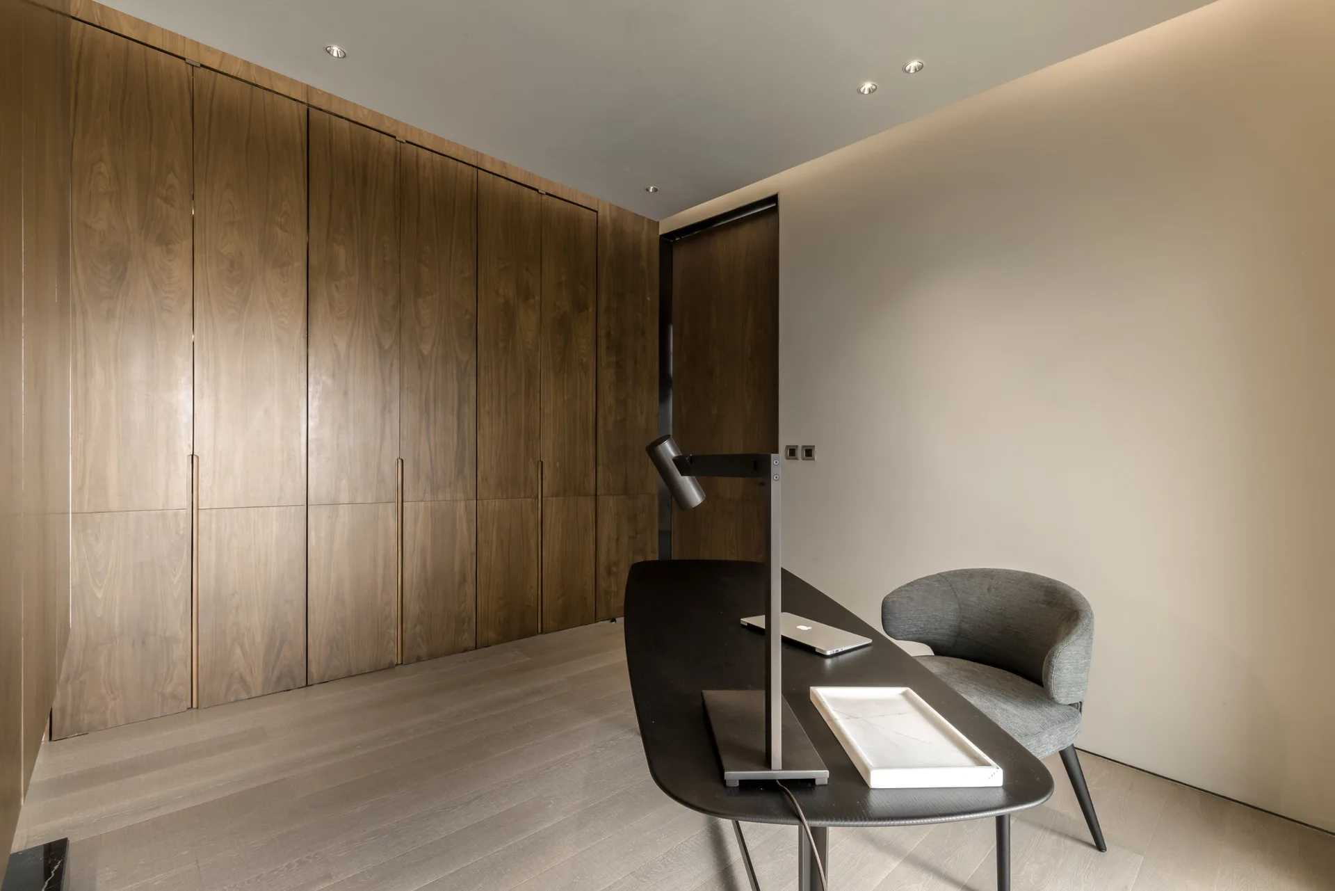 A modern home office with a dark curved desk, grey armchair, and a full wall of floor-to-ceiling wooden cabinets with integrated handles.