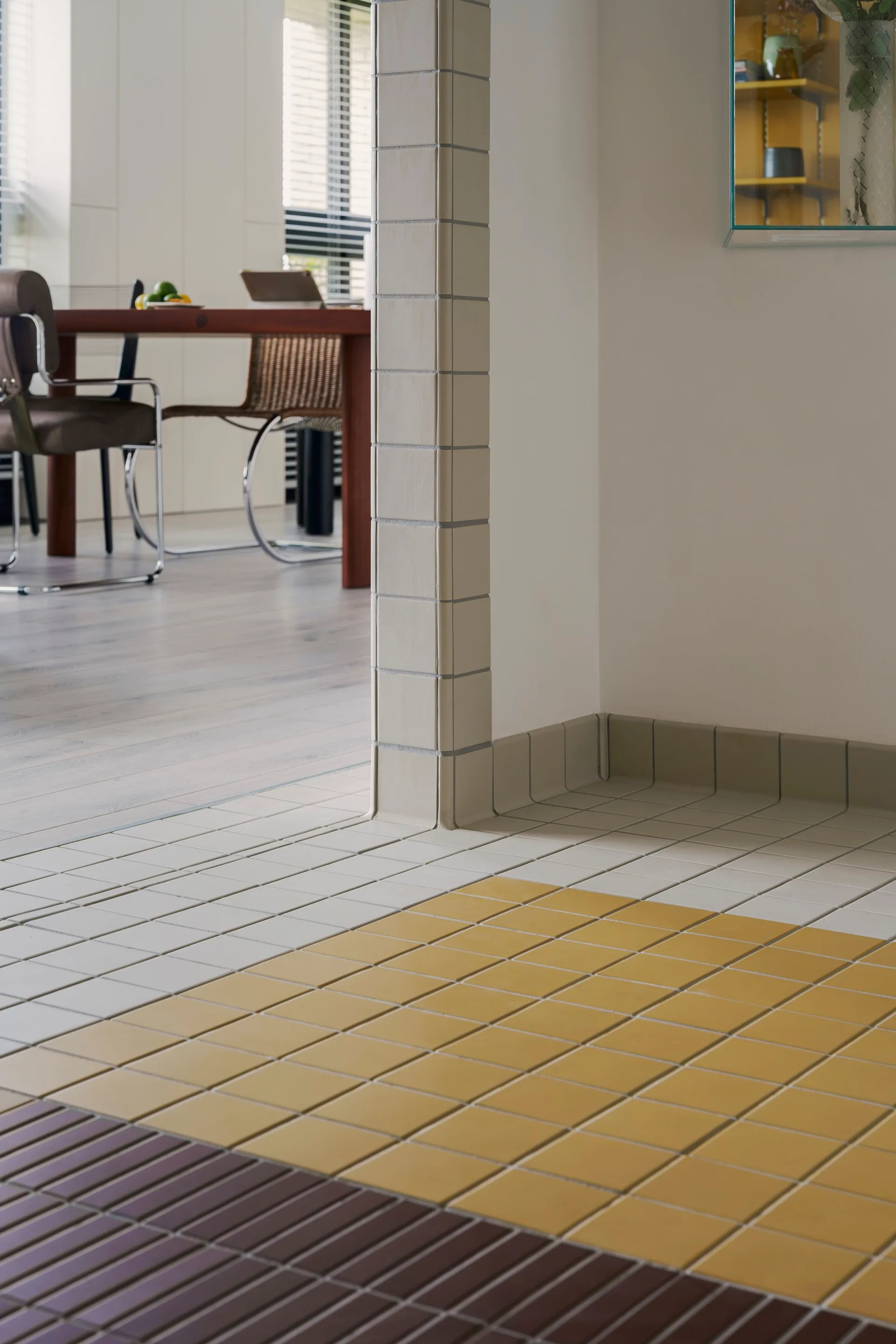 A close-up view of a transition area featuring yellow, brown, and white floor tiles that meet a tiled wall column with a dining room in the background.