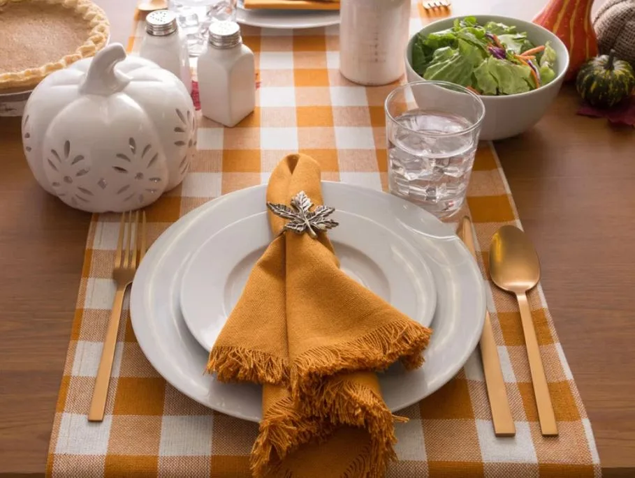 A fall table setting features white plates, a fringed mustard yellow napkin with a leaf ring, gold flatware, and an orange gingham runner.