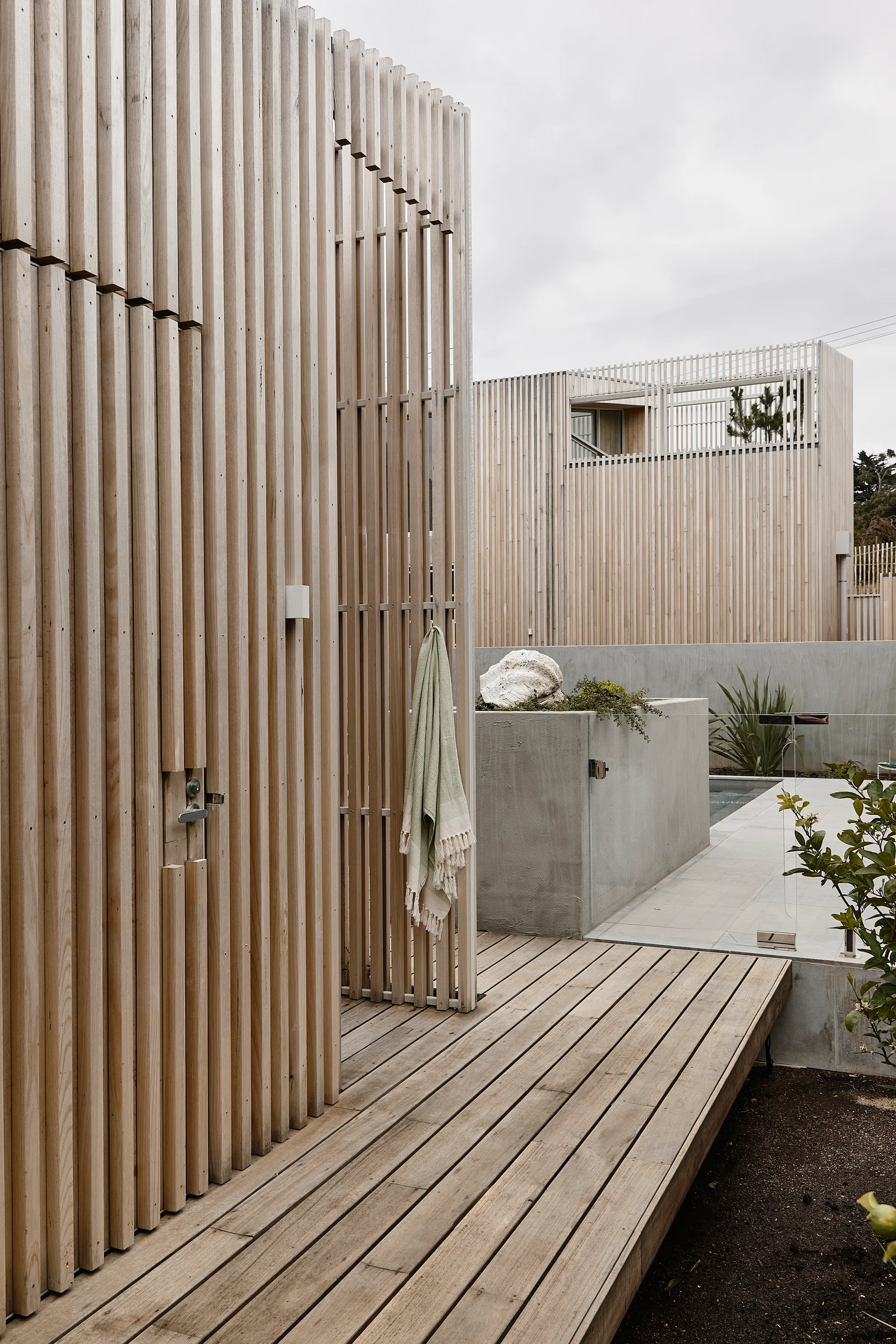 A modern outdoor deck features vertical light-toned wood slat walls, a matching wood plank floor, and a smooth concrete planter near a pool.
