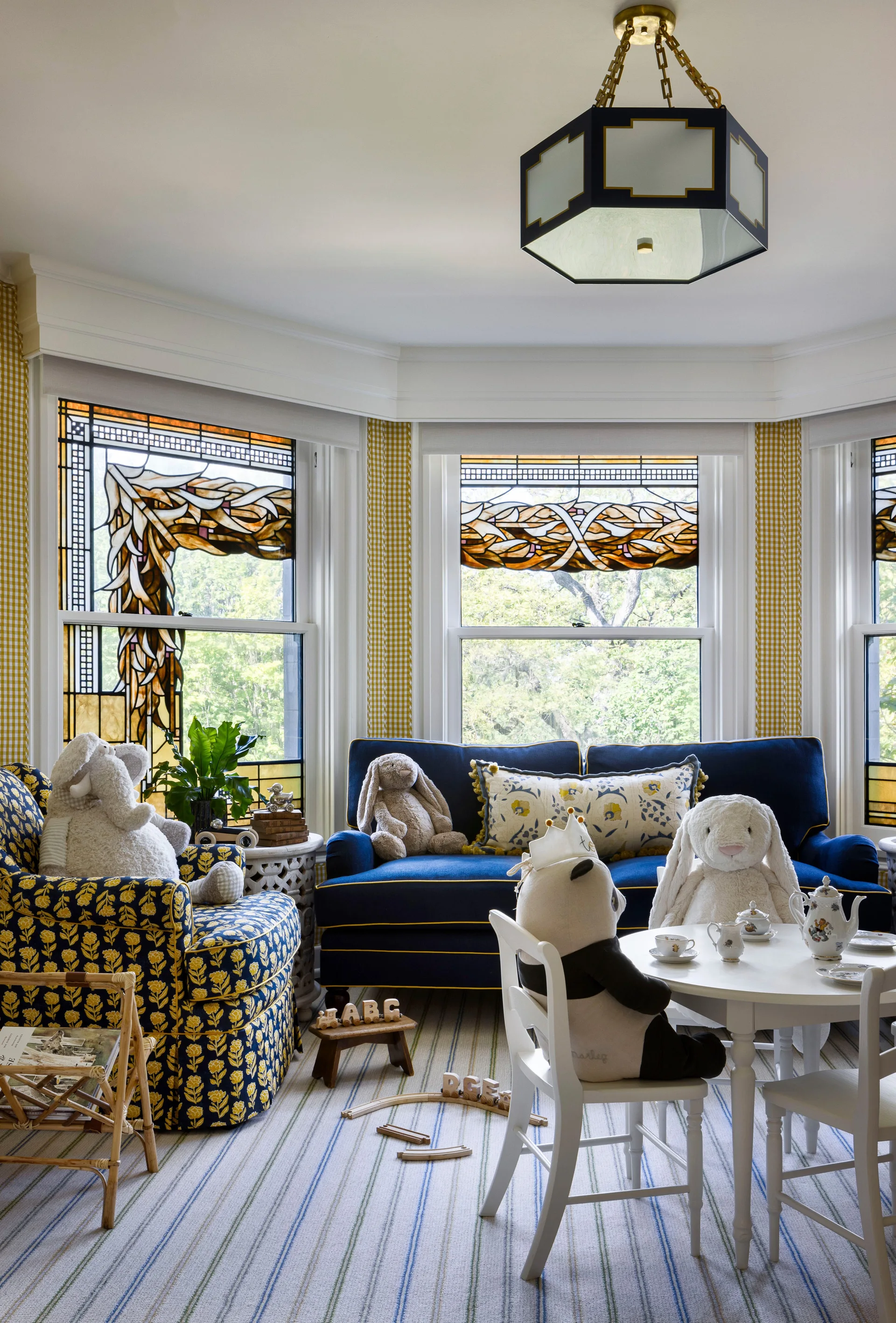 A kids play area features a navy sofa inside a stained-glass bay window next to a white activity table set with stuffed animals.