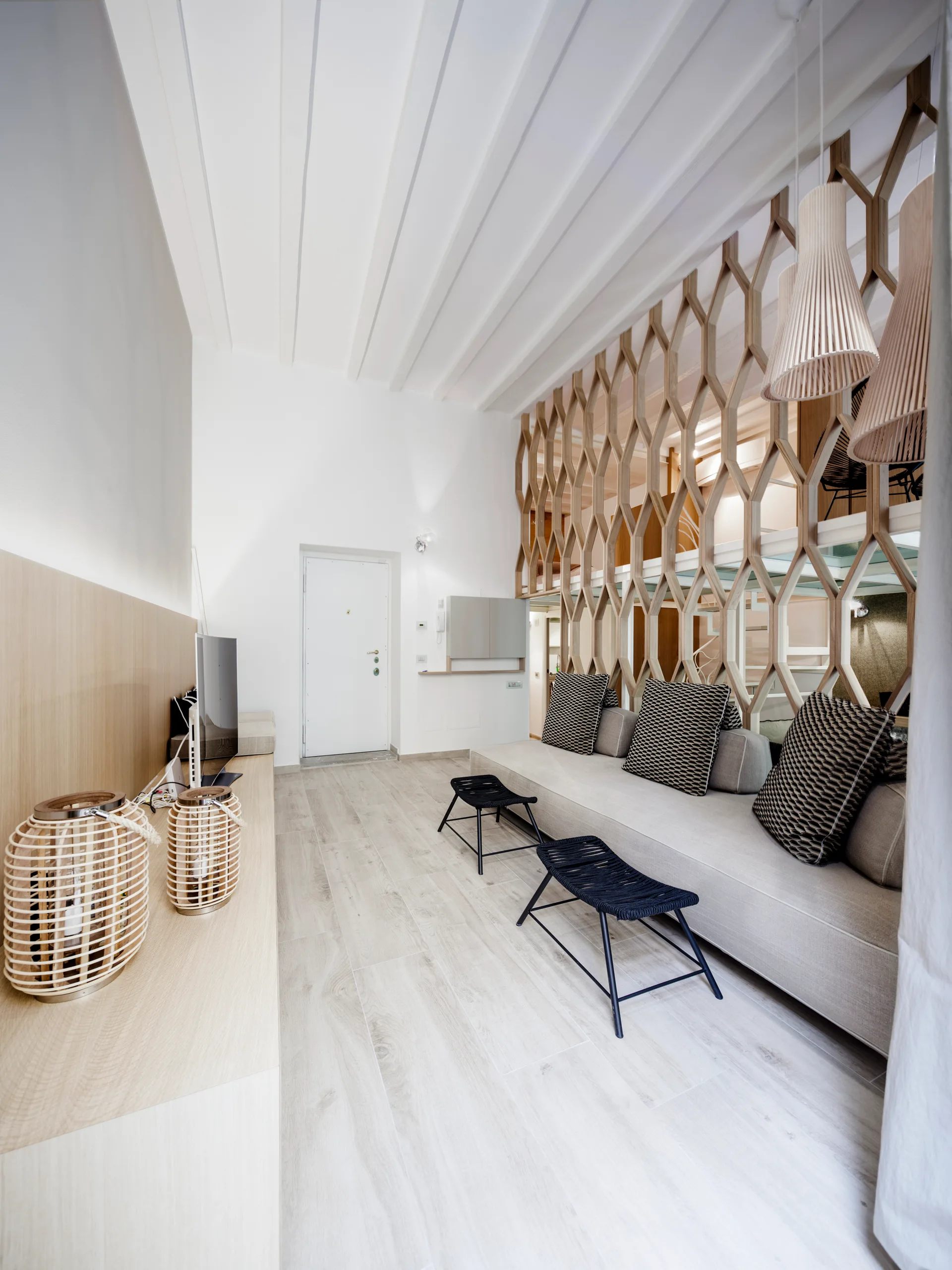 A modern living room features a long beige sofa beneath a geometric wooden lattice partition wall, with light wood floors and two black stools.
