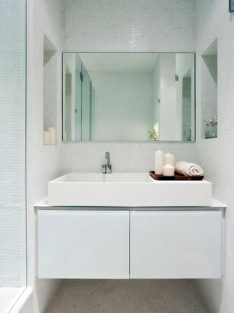 A wall-mounted white vanity with a rectangular sink and a frameless mirror is set against walls covered in small white mosaic tiles.
