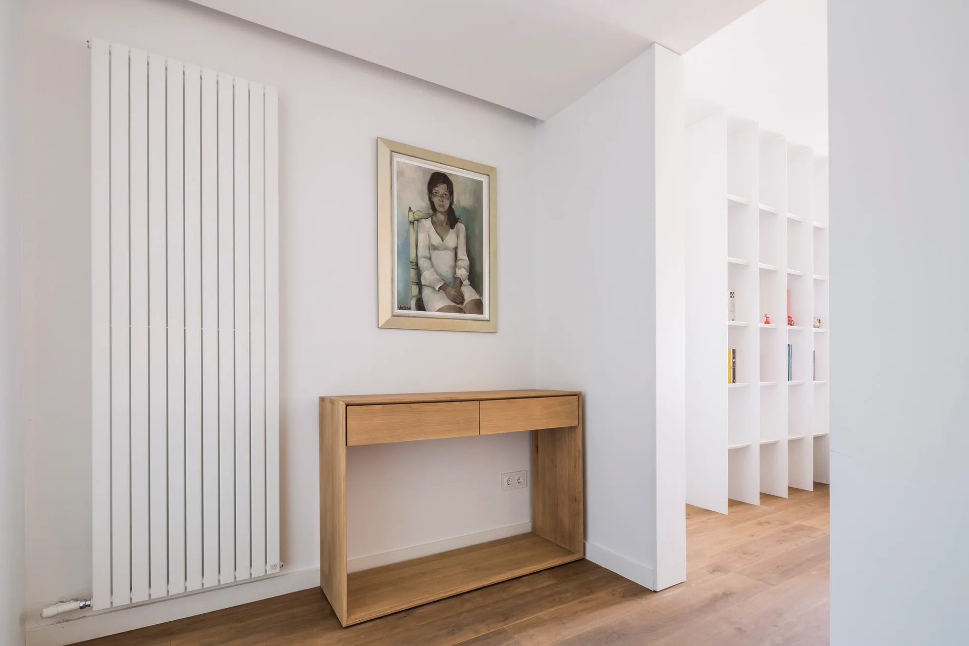 A minimalist hallway features a natural wood console table under a framed portrait, next to a tall white vertical wall radiator and built-in shelving.