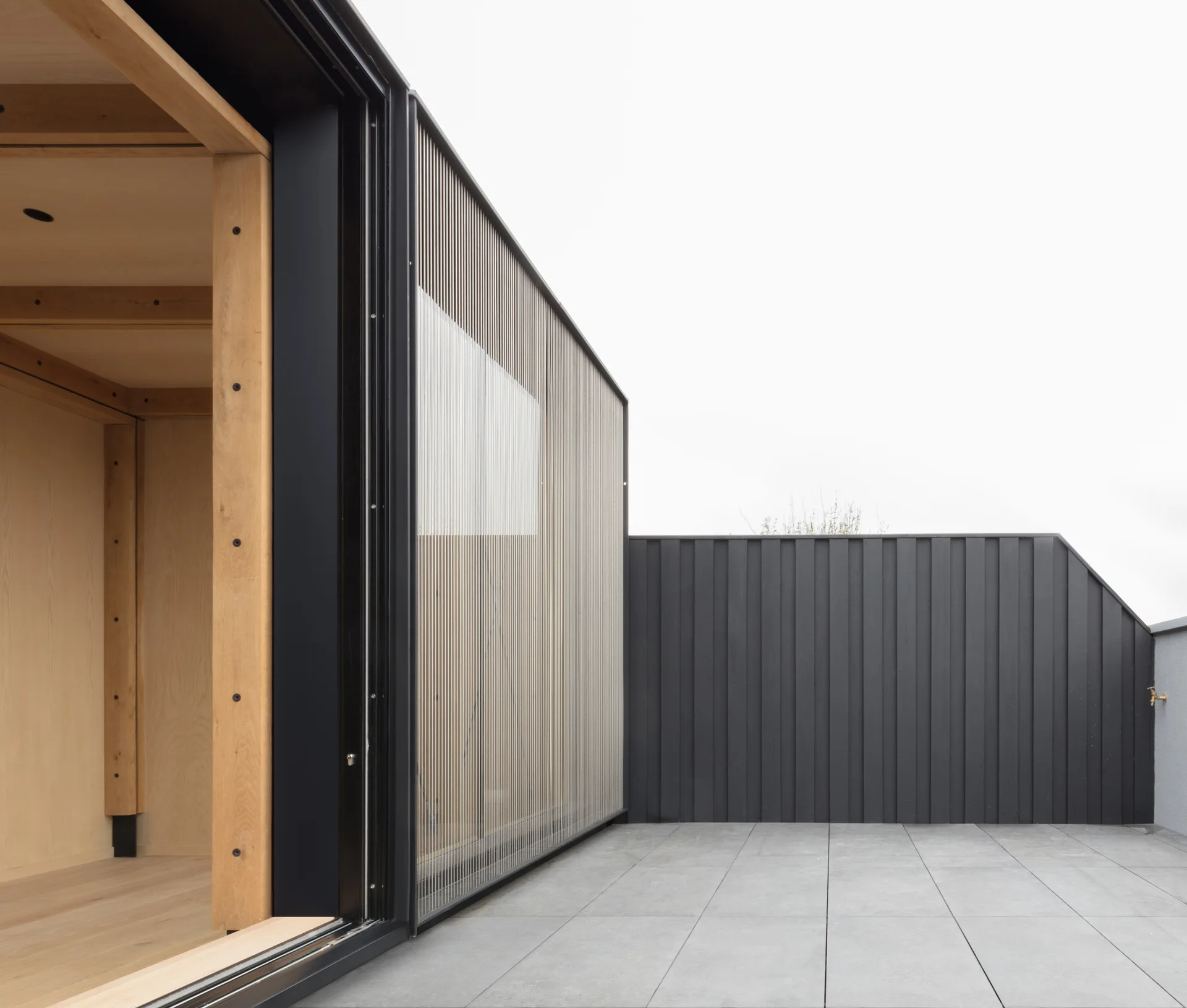 A minimalist outdoor terrace features grey tiled flooring, dark vertical wood cladding, and a slatted metal screen next to a sliding glass entryway.