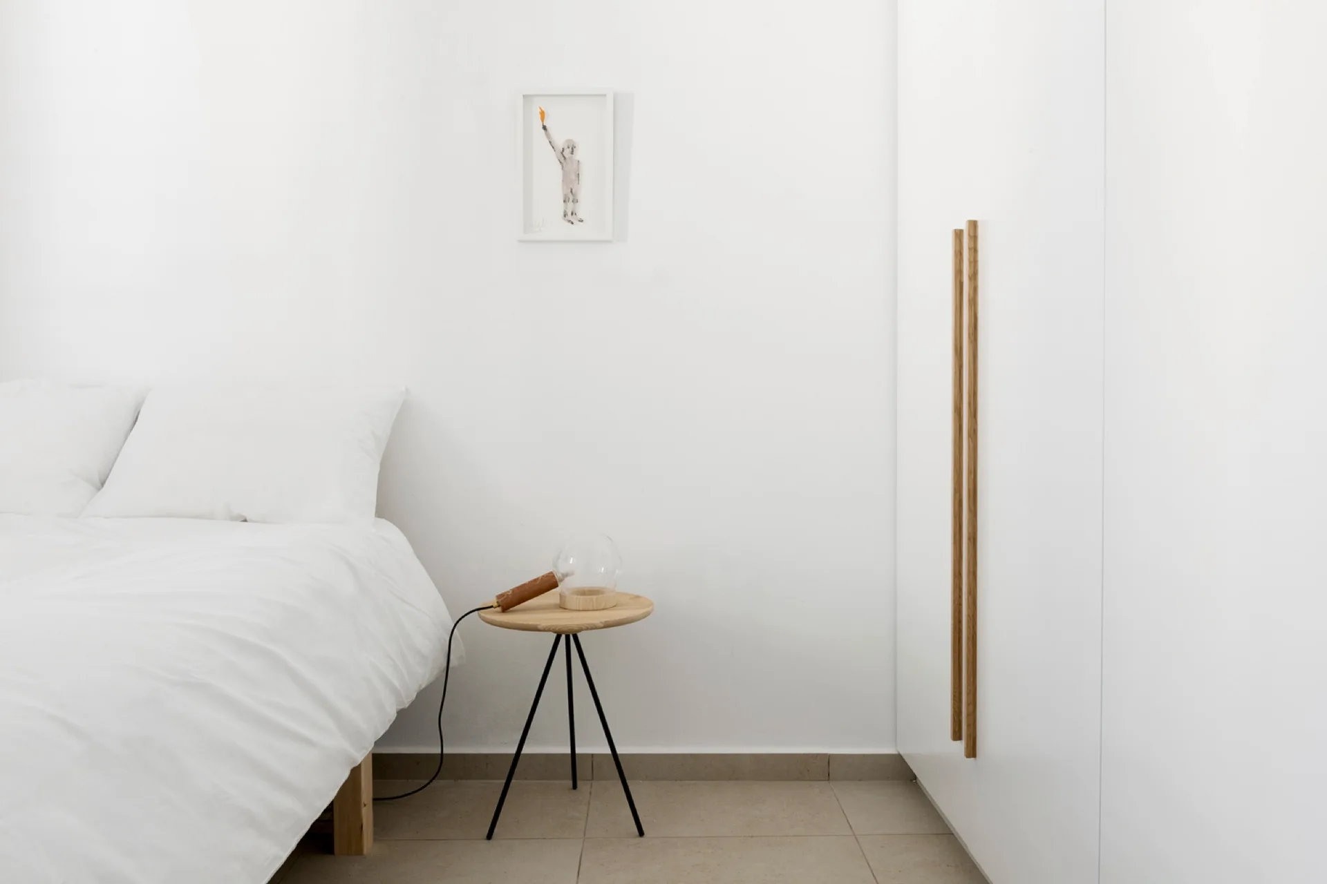A minimalist white bedroom featuring a bed with white linens, a small round wooden nightstand, and a white built-in wardrobe with long wooden handles.