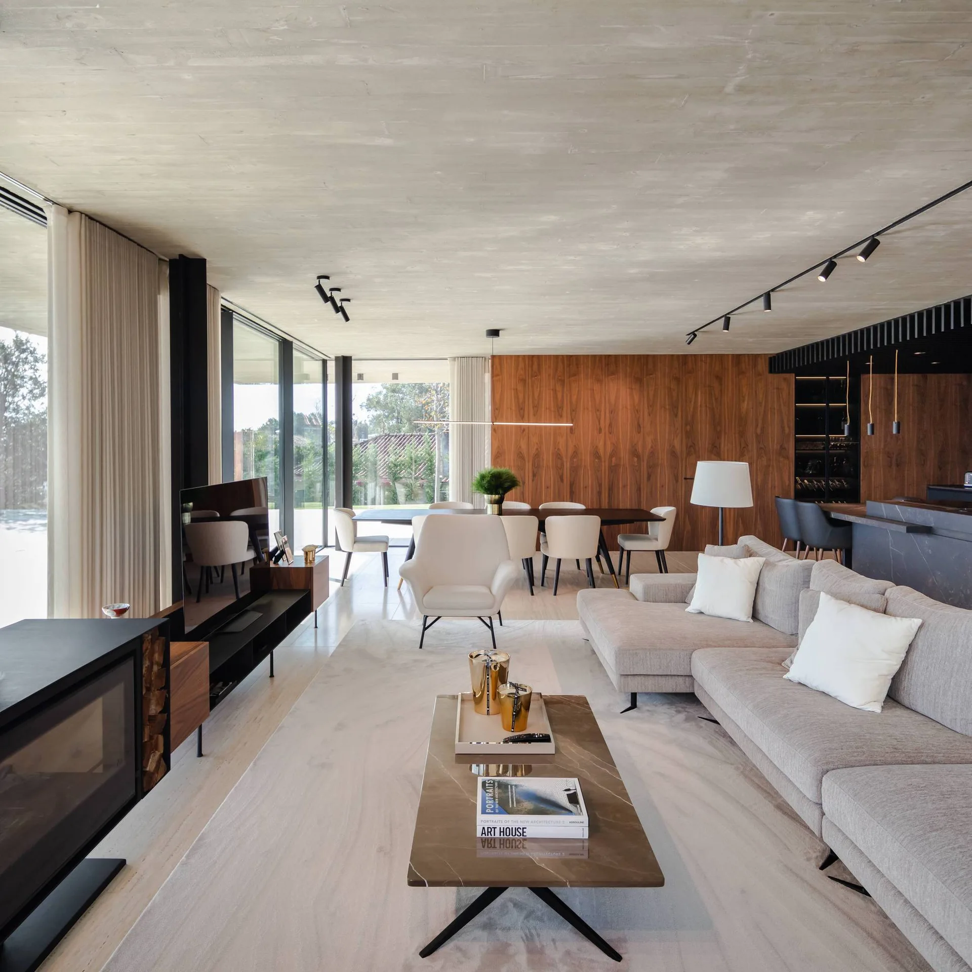 A modern open-plan living room features a grey sectional sofa, a stone-top coffee table, a concrete ceiling, and a large walnut wood paneled wall.
