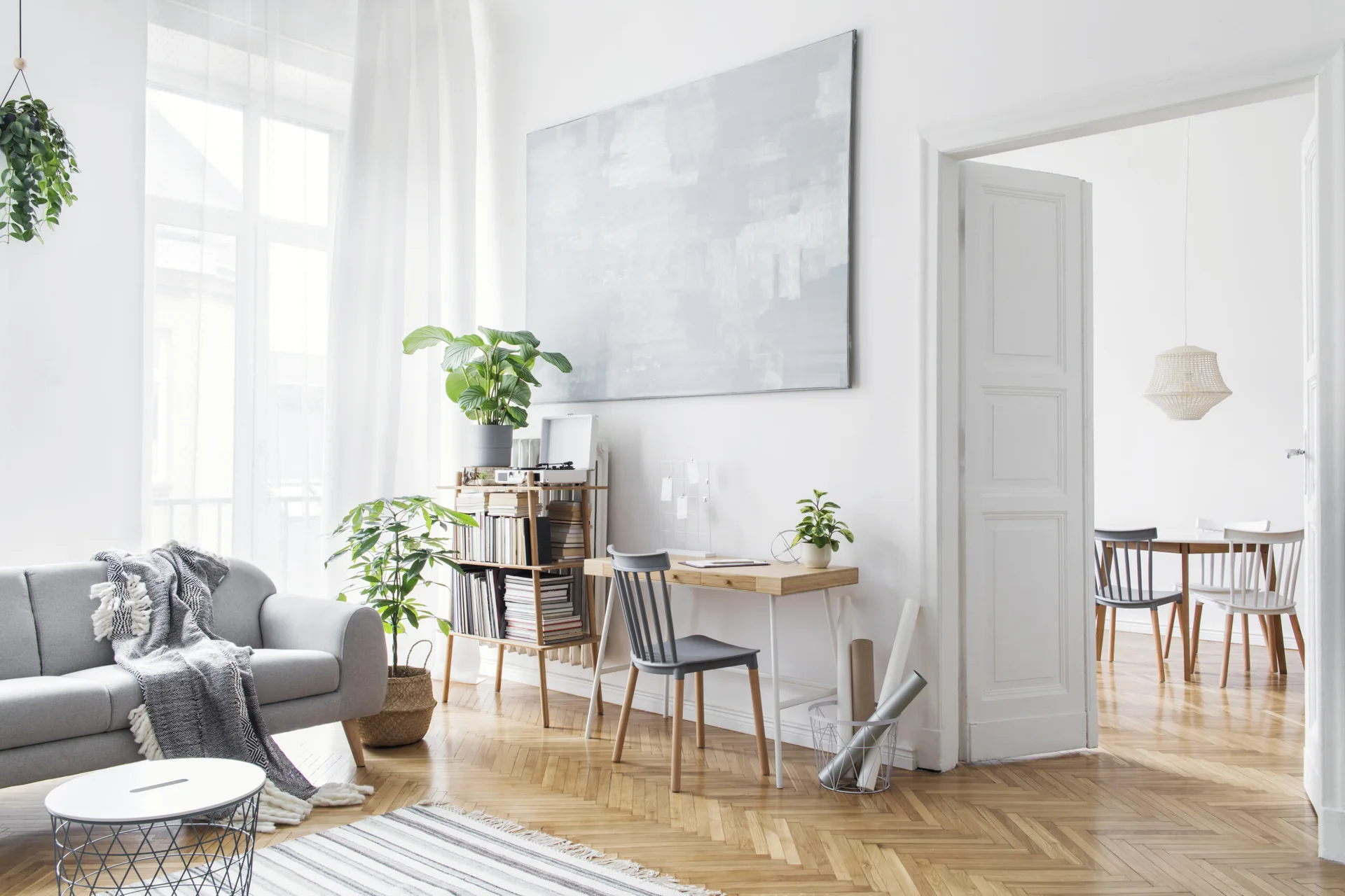 A bright white Scandinavian living room features a grey sofa, a wooden desk with a spindle chair, chevron parquet flooring, and a large abstract canvas.