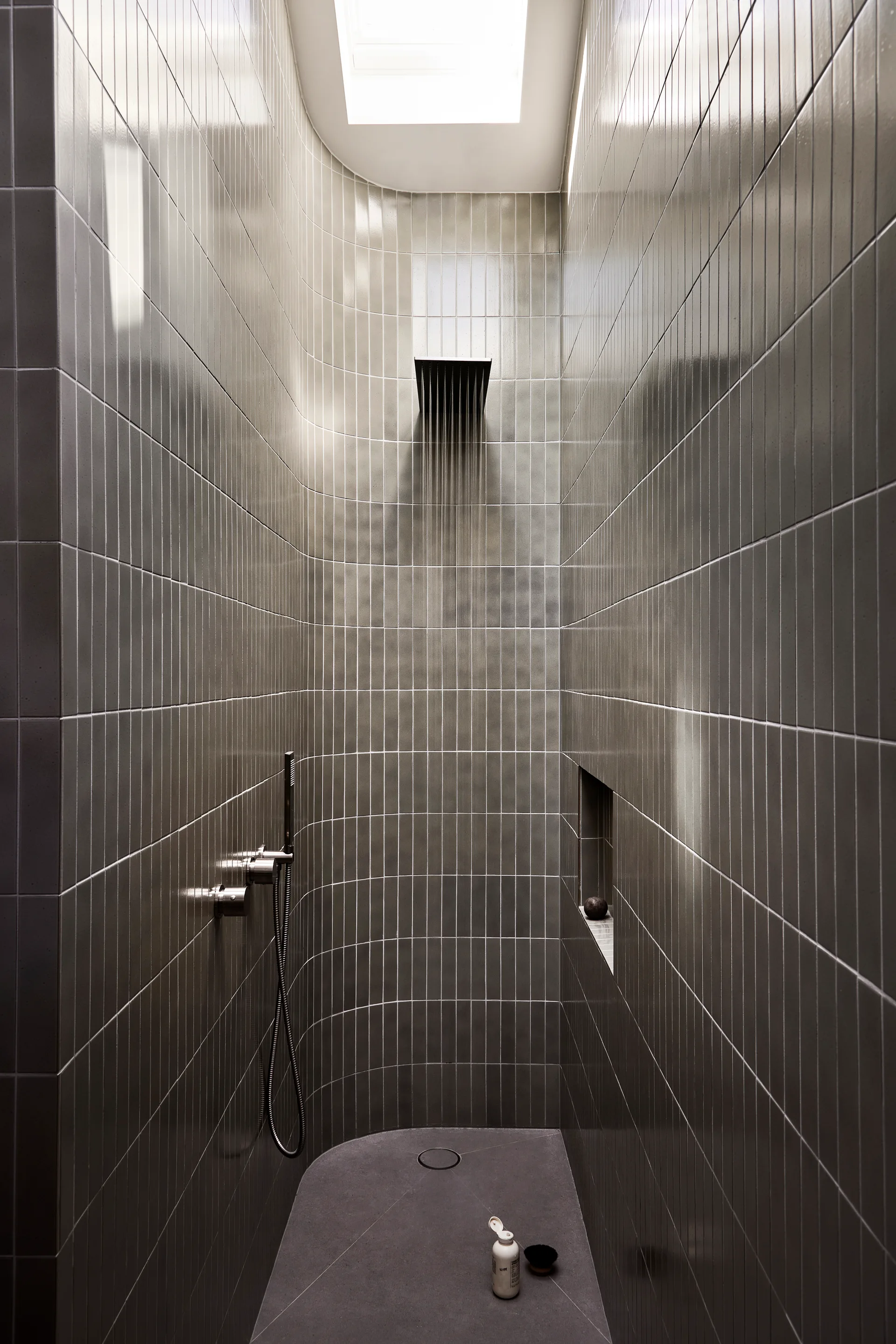 A narrow gray tiled shower features vertically stacked ceramic tiles, a curved back wall, a recessed wall niche, and a bright skylight.
