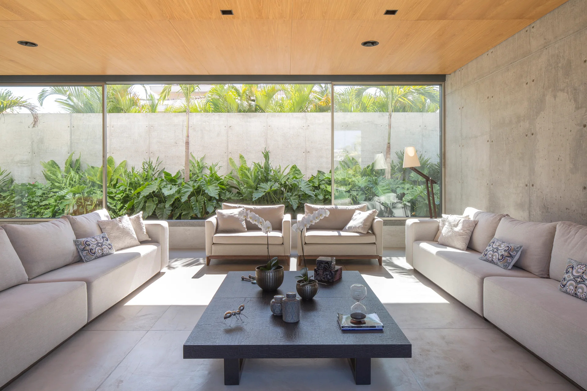 A modern living room features cream sofas, a black coffee table, an exposed concrete wall, a wood ceiling, and a window facing a leafy garden.