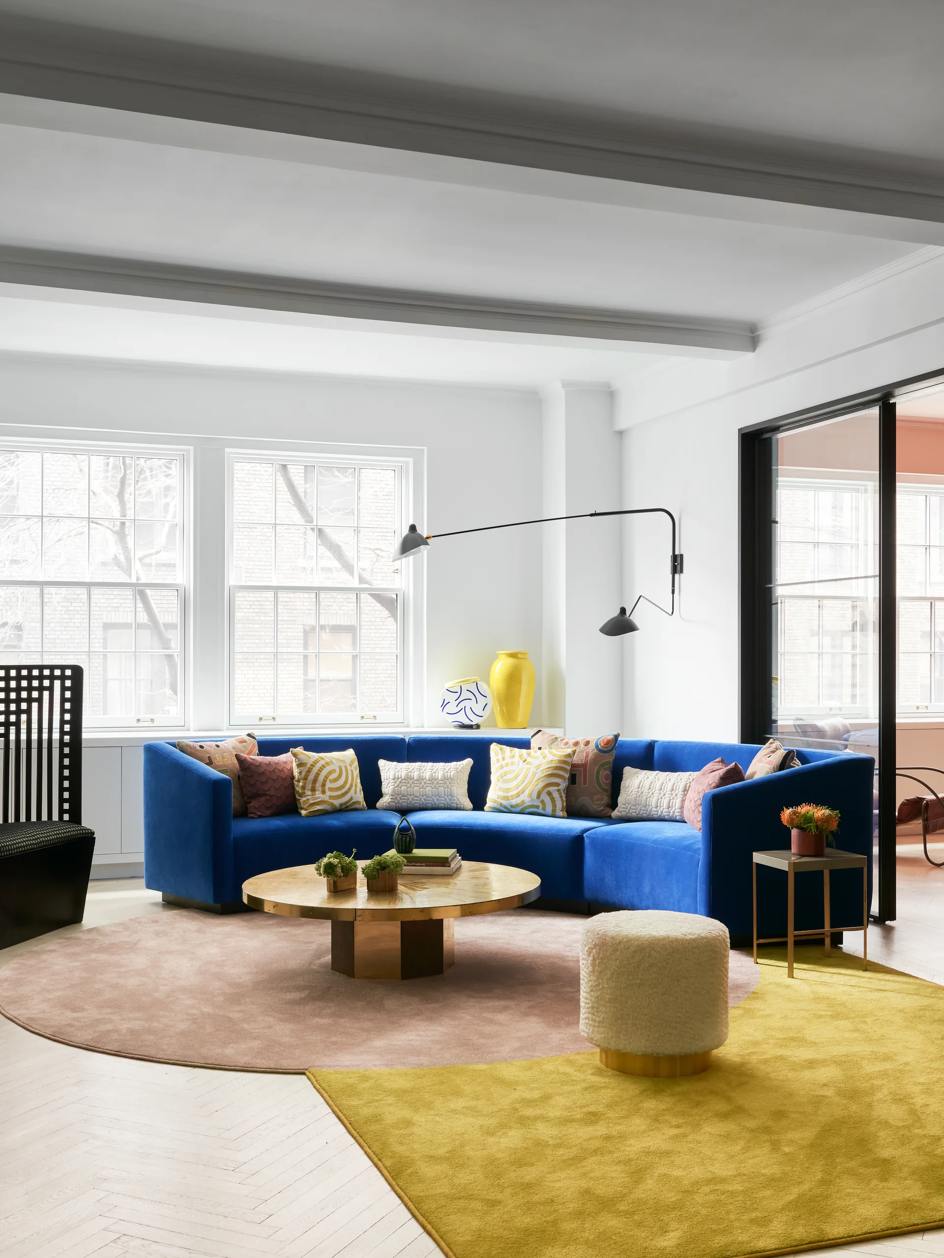 A contemporary living room features a bright blue curved velvet sofa, a round gold coffee table, and overlapping pink and yellow rugs.