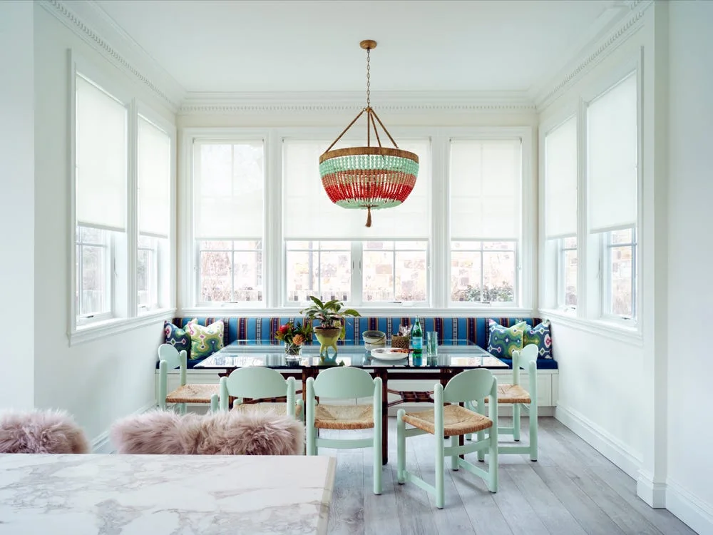 A bright dining nook features mint green chairs and a striped banquette under a colorful beaded chandelier surrounded by windows.