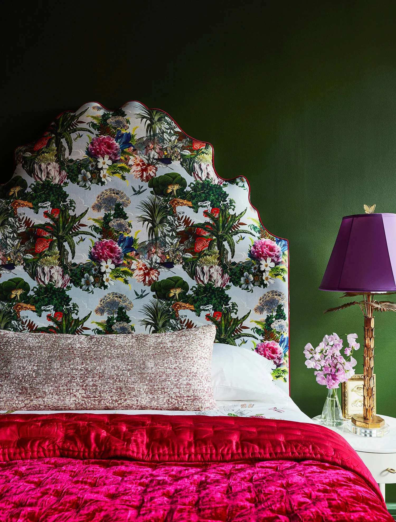 A bedroom featuring a scalloped floral headboard against an olive green wall, a fuchsia velvet quilt, and a brass palm-shaped lamp with a purple shade.