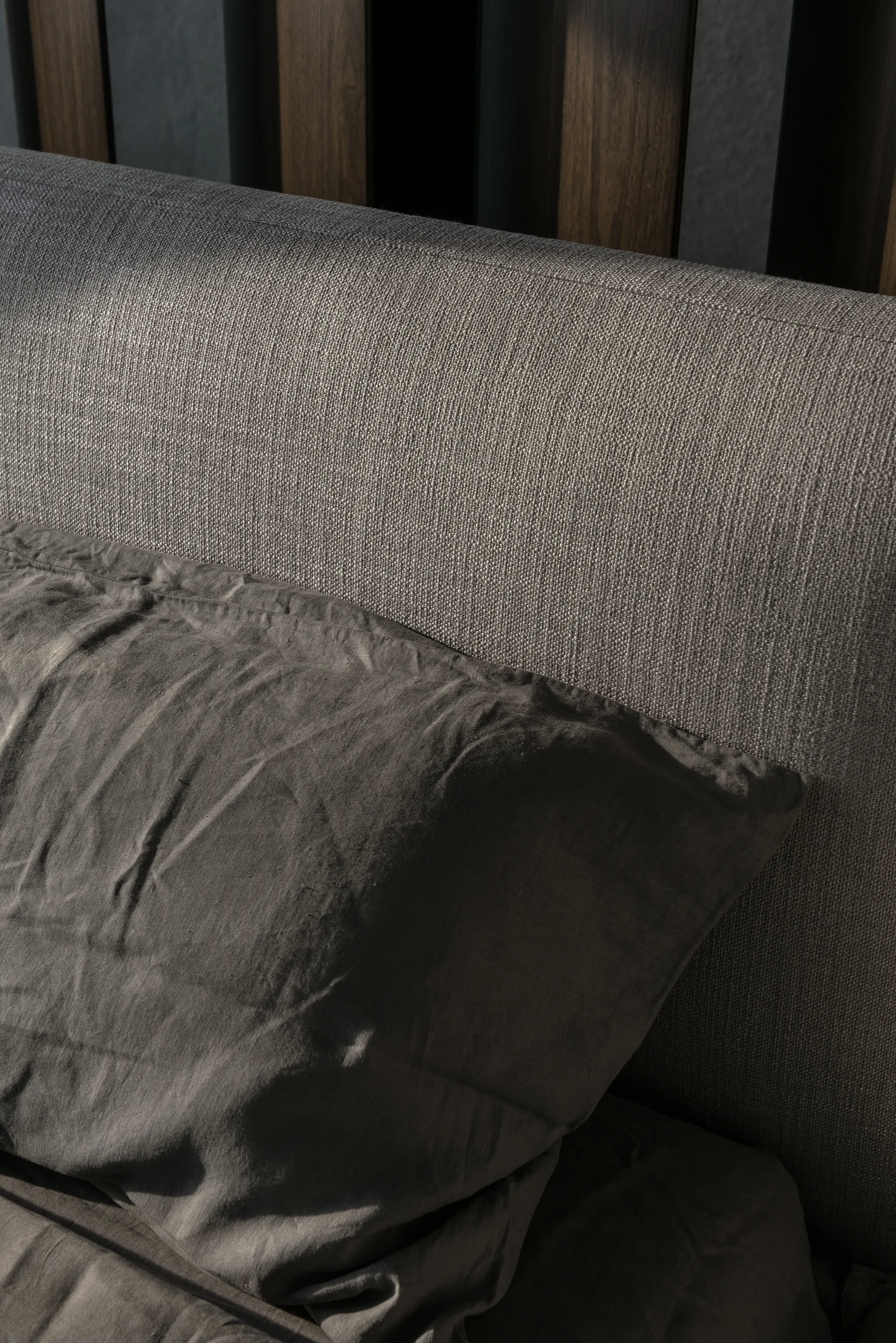 A close-up of a textured grey fabric headboard and dark grey pillow set against a wall treatment of vertical wood slats.