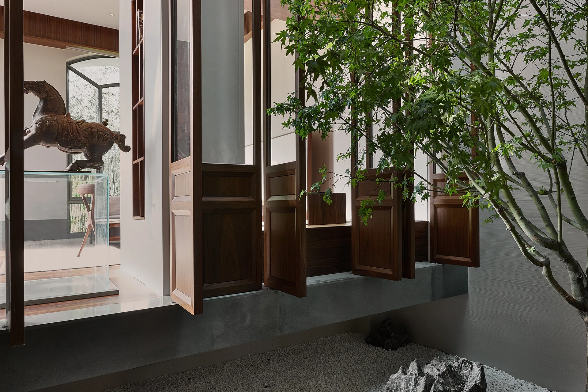 An indoor courtyard features hinged wooden screens on a concrete ledge next to a gravel garden with a small green tree and ornamental rock.