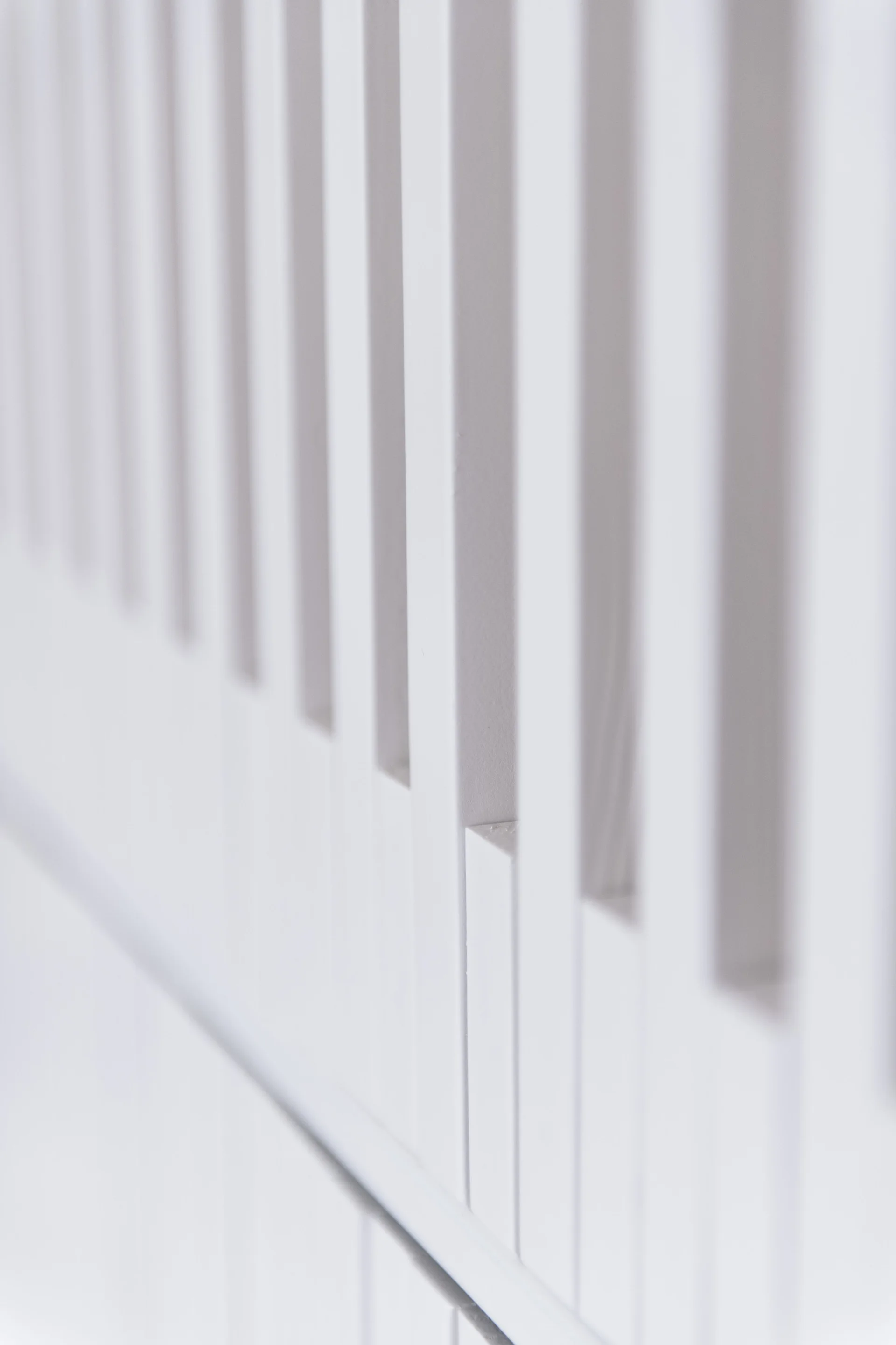 A close-up perspective of a matte white panel featuring closely spaced vertical wooden slats that create a rhythmic shadow pattern.