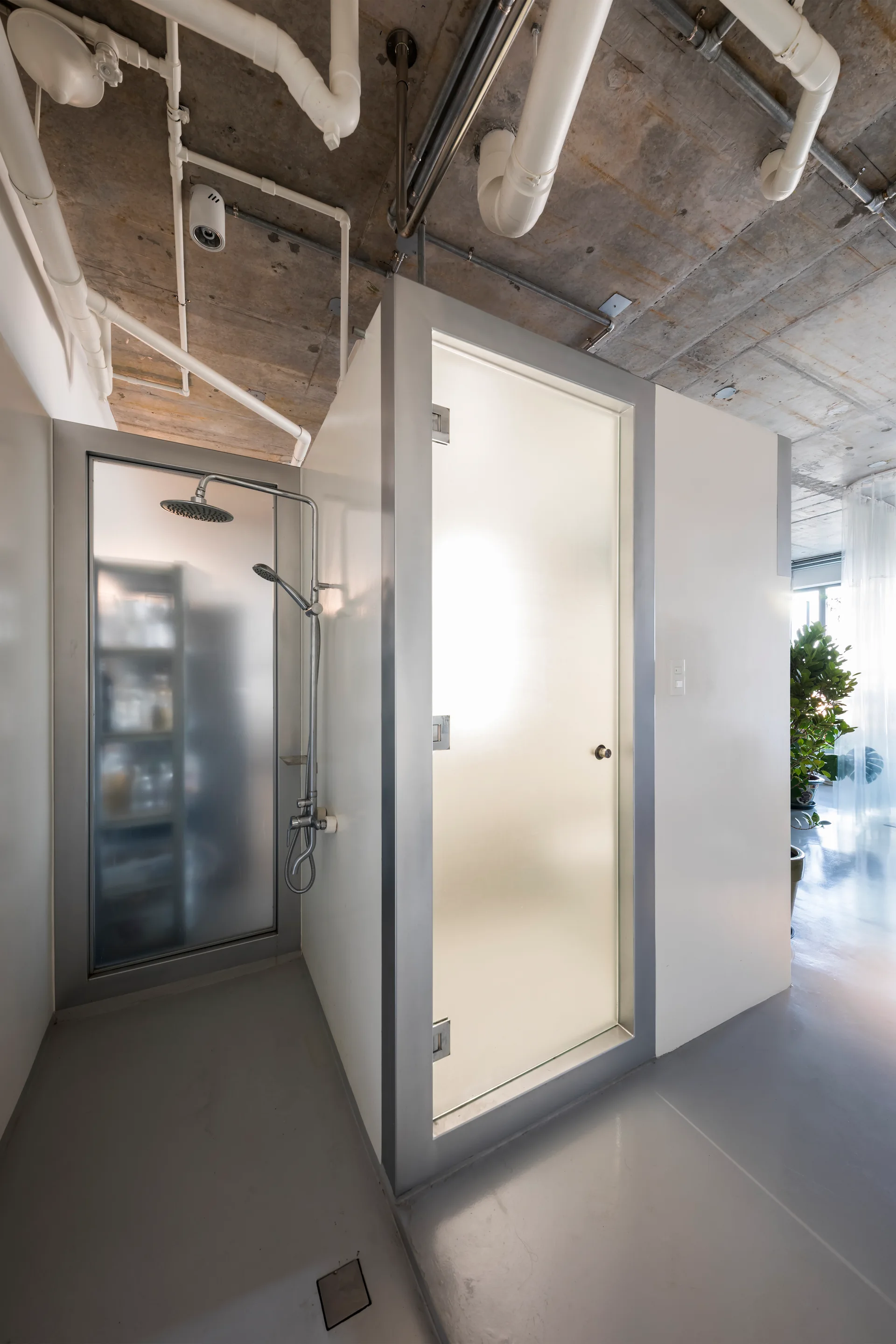 A modern white shower stall with a frosted glass door stands beneath an exposed concrete ceiling with white pipes in an industrial bathroom.
