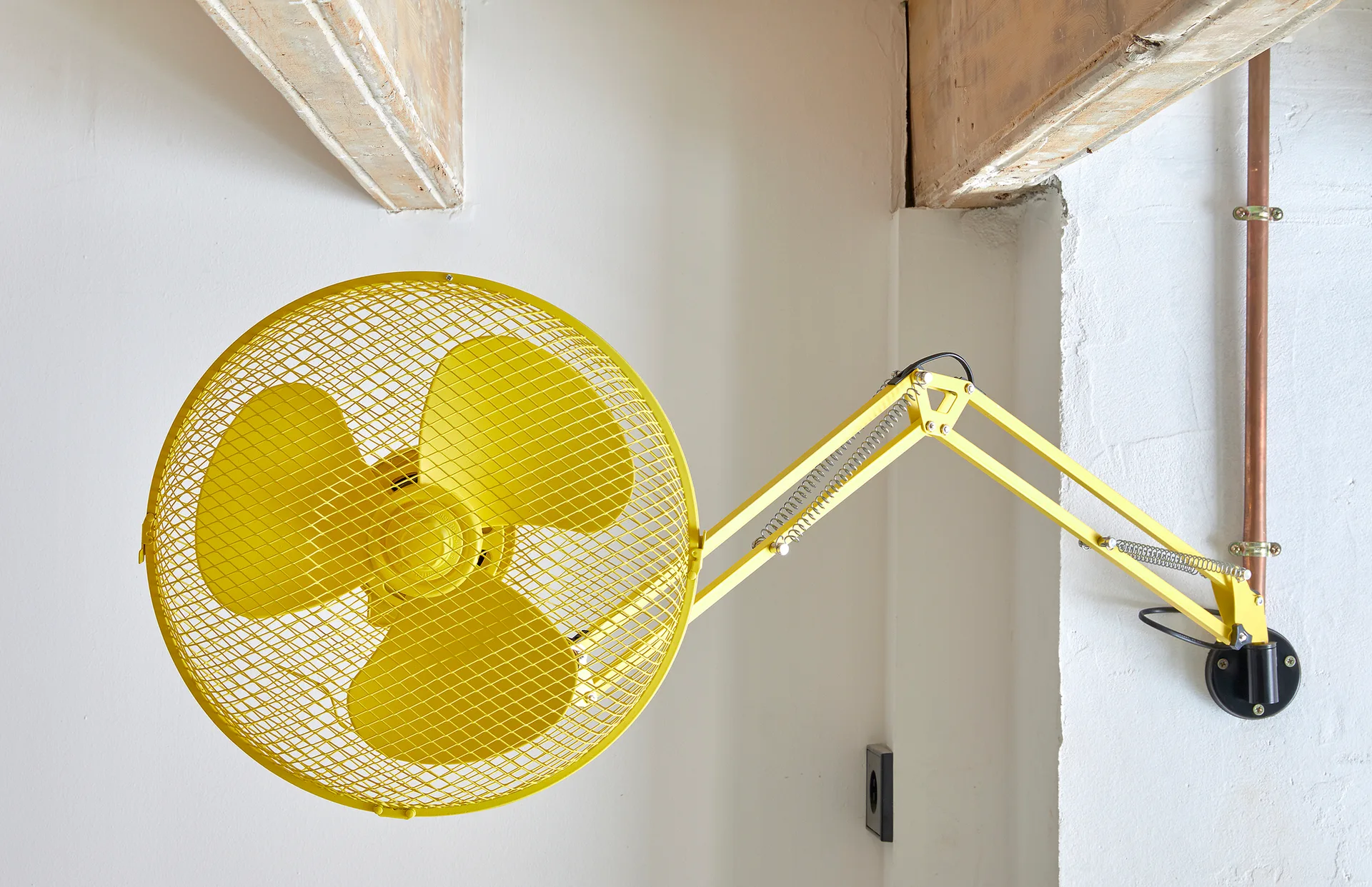 A bright yellow industrial wall fan with an articulating arm is mounted on a white wall below exposed wooden ceiling beams.