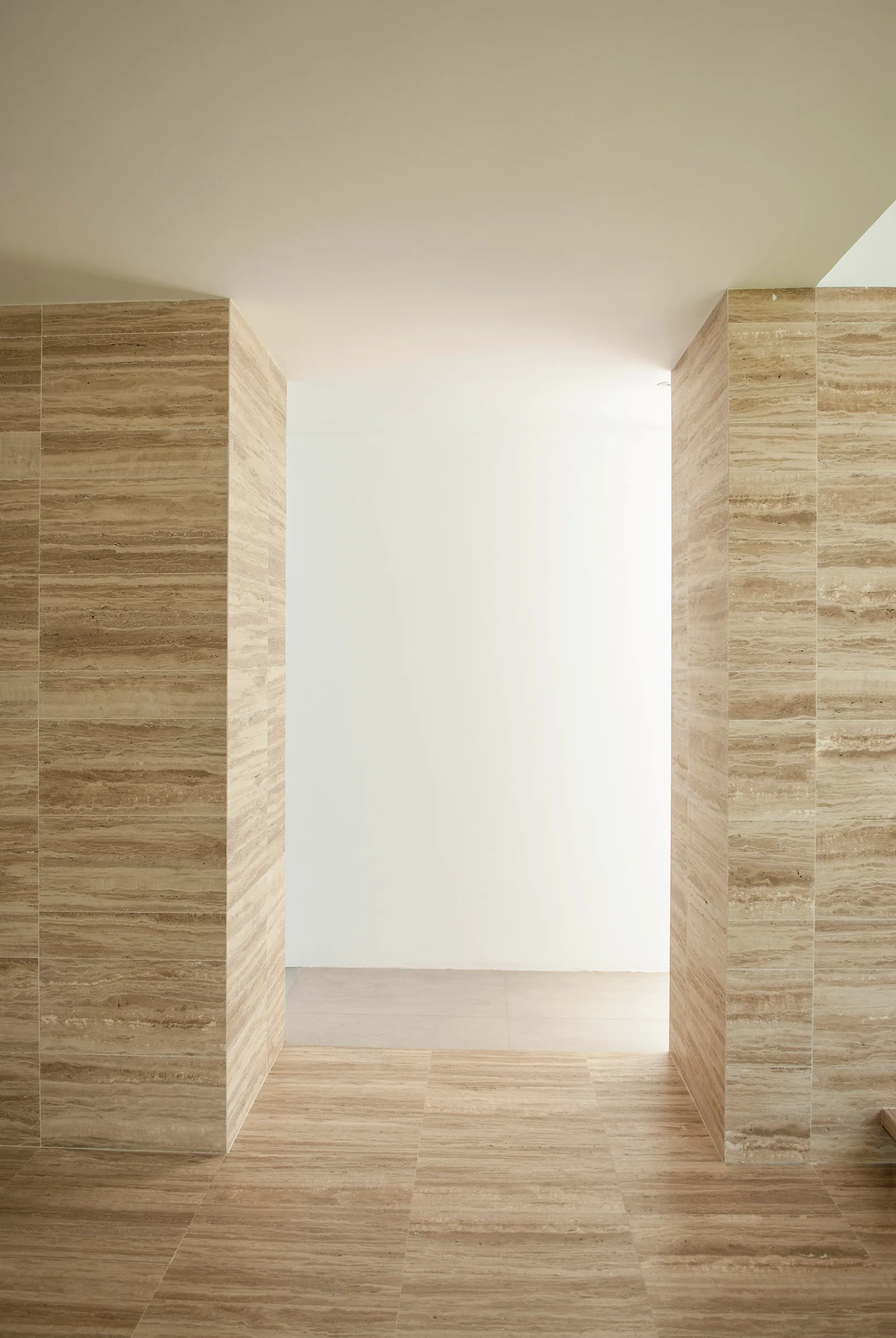 A minimalist interior passage framed by two beige travertine stone pillars with horizontal banding, leading to a brightly lit white hallway.