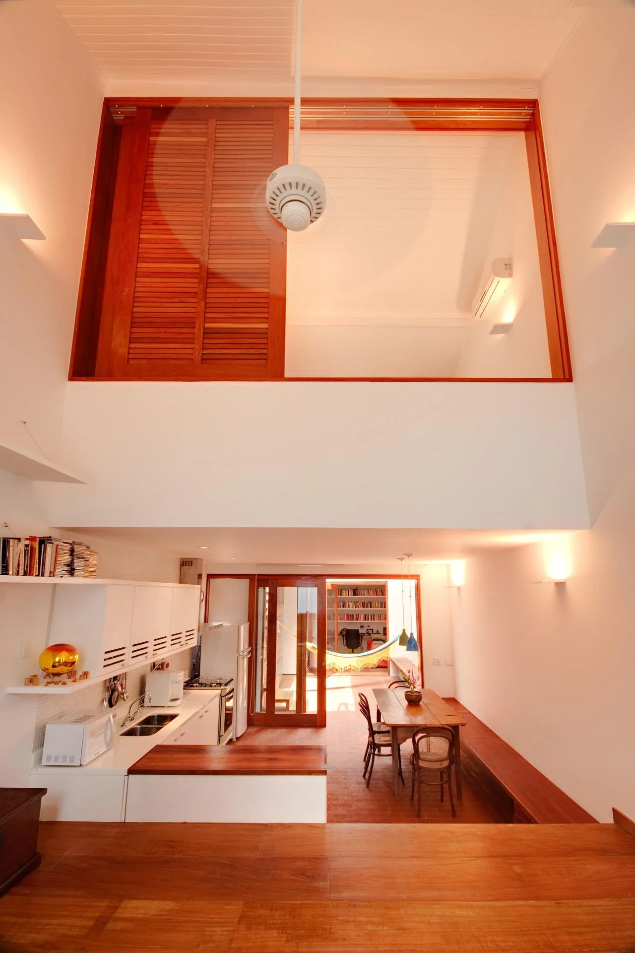 A double-height kitchen and dining area features white cabinetry, brick flooring, a wooden dining table, and a high mezzanine with a sliding wood shutter.