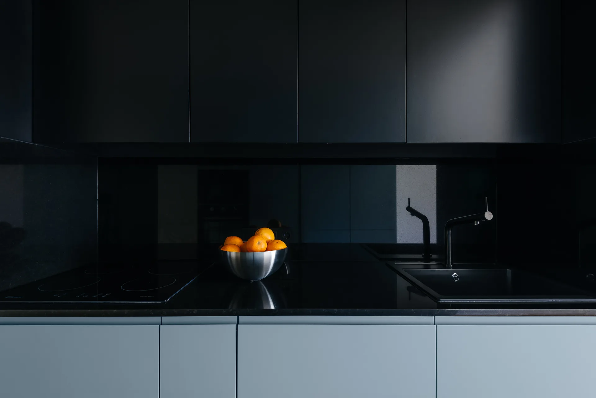 A minimalist kitchen workspace shows matte black upper cabinets, a dark countertop with a sink and cooktop, and a metal bowl of oranges.