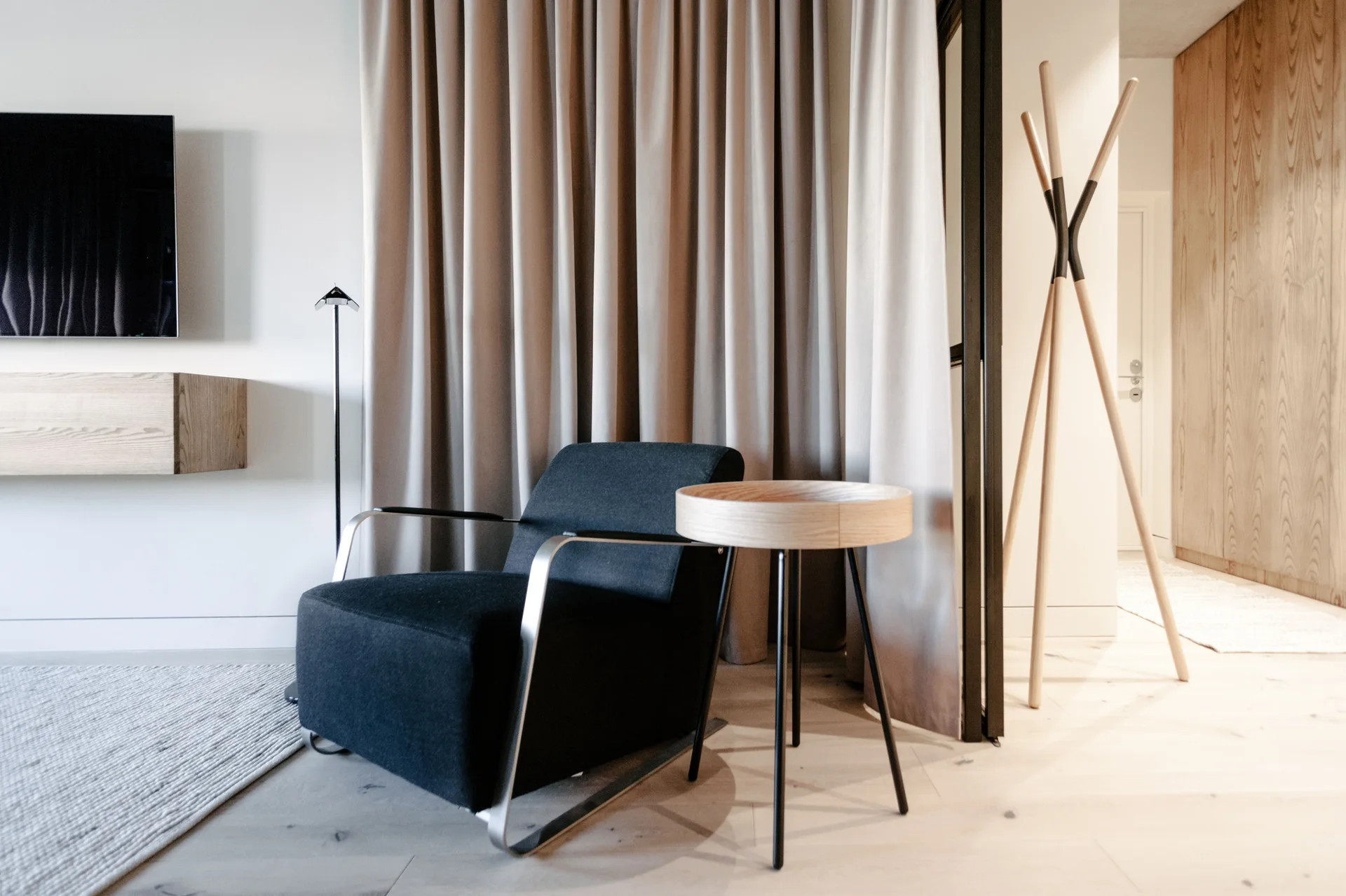 A dark fabric armchair sits next to a round wooden side table against floor-to-ceiling beige curtains in a modern room.