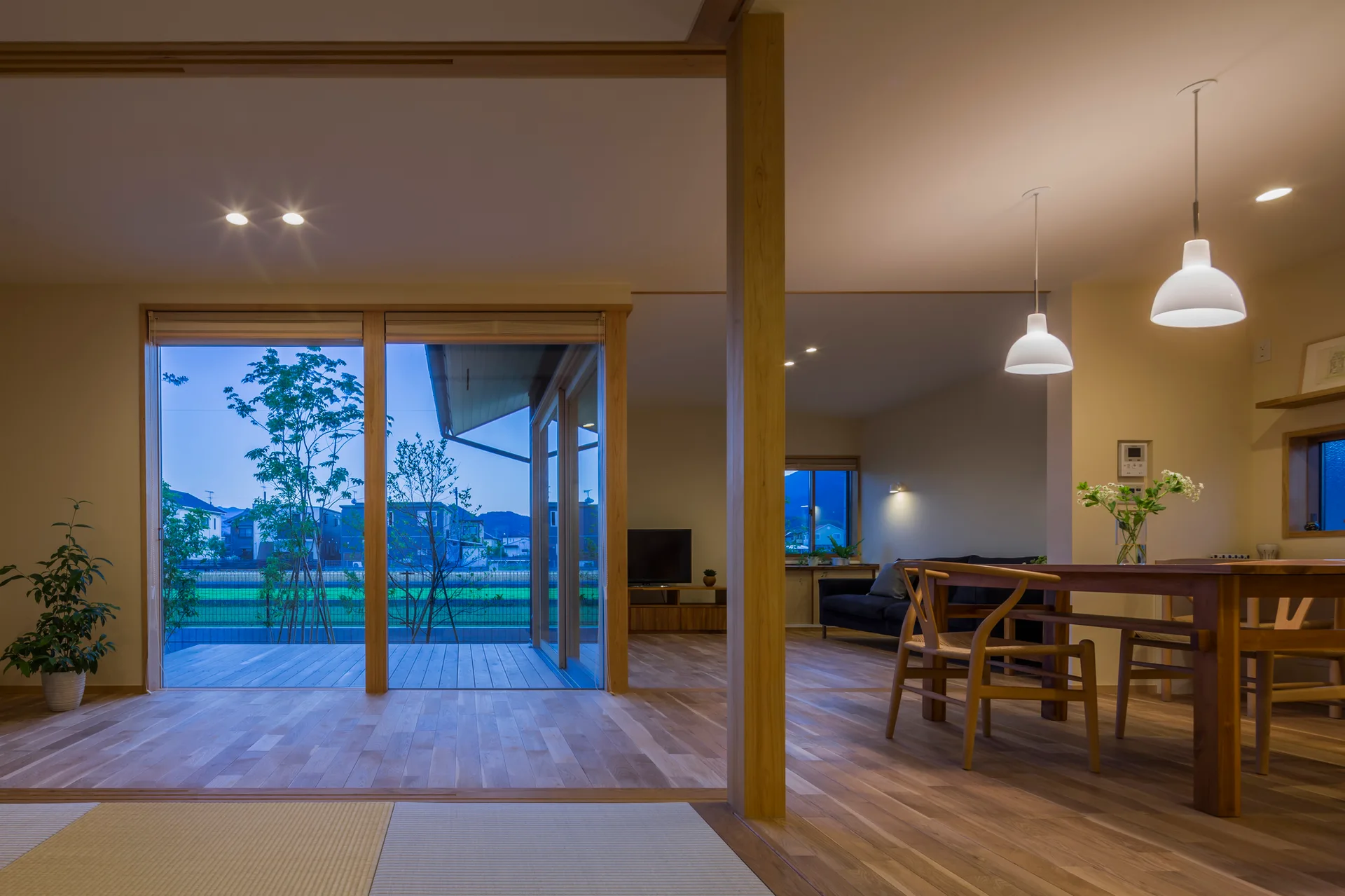 A modern open-plan home interior features wood floors, a dining table with chairs under pendant lights, and sliding glass doors opening to a deck.