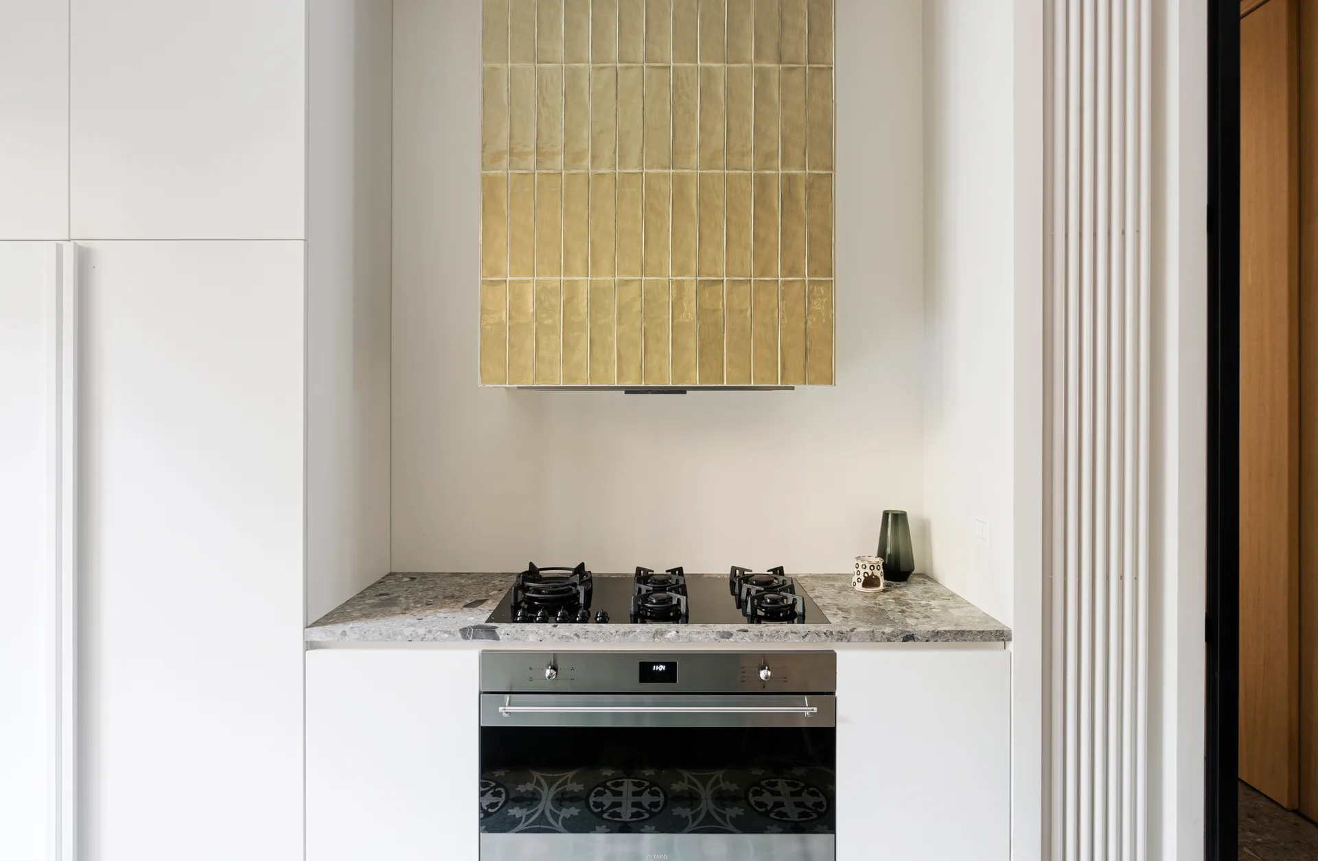 A kitchen niche features white cabinets, a grey speckled countertop with a gas cooktop, and a range hood covered in vertical gold tiles.