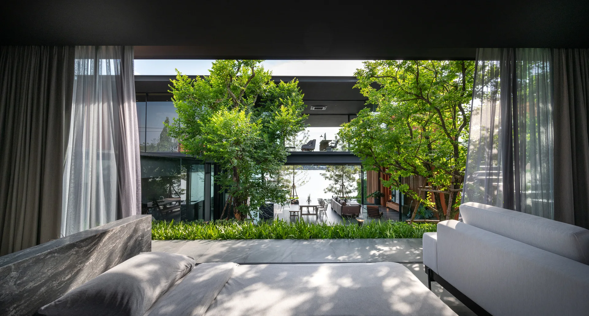 A view from a minimalist bedroom looking out through expansive glass windows onto a lush, tree-filled courtyard with outdoor furniture.