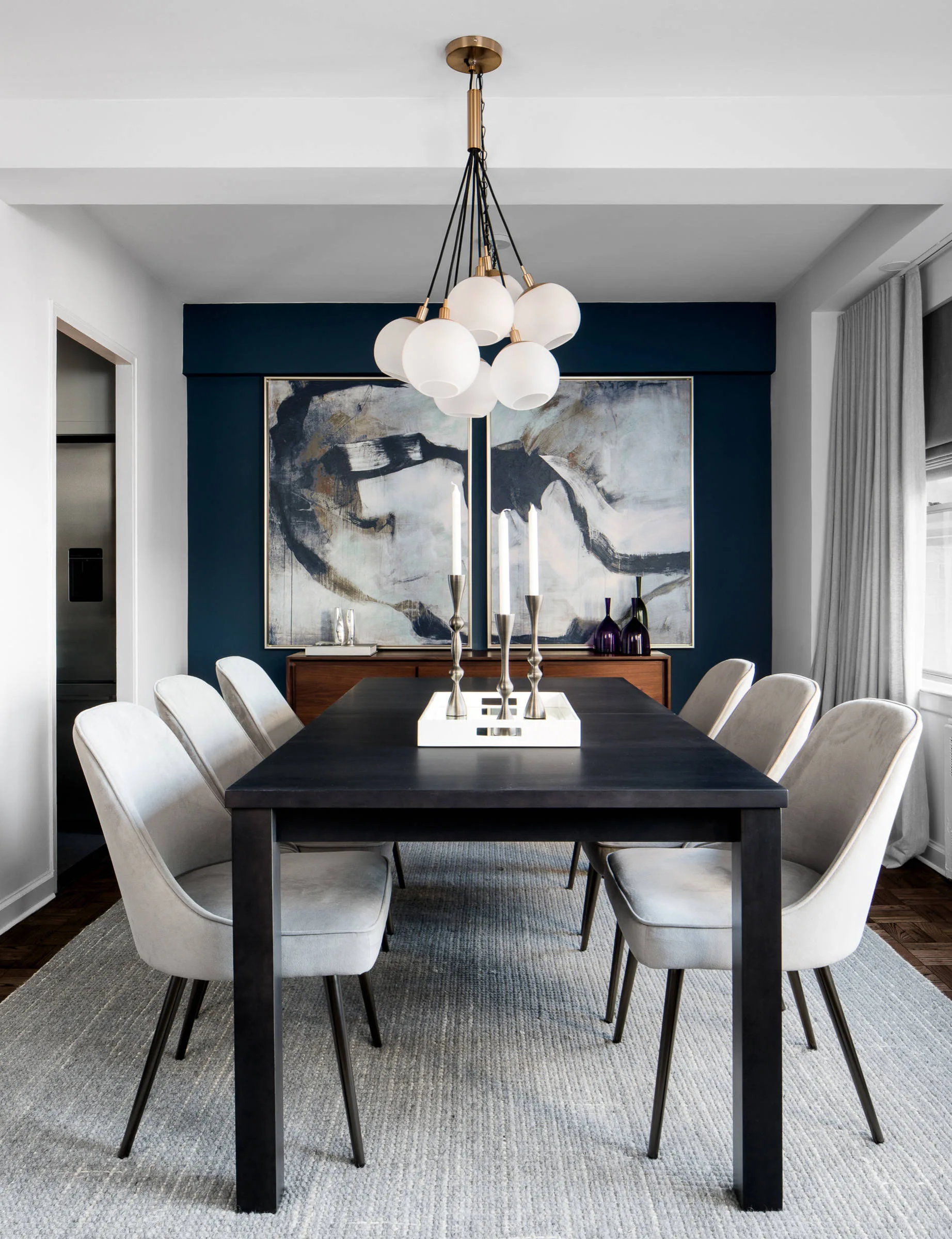 A modern dining room features a black wooden table and light grey upholstered chairs set under a multi-globe chandelier against a navy accent wall.