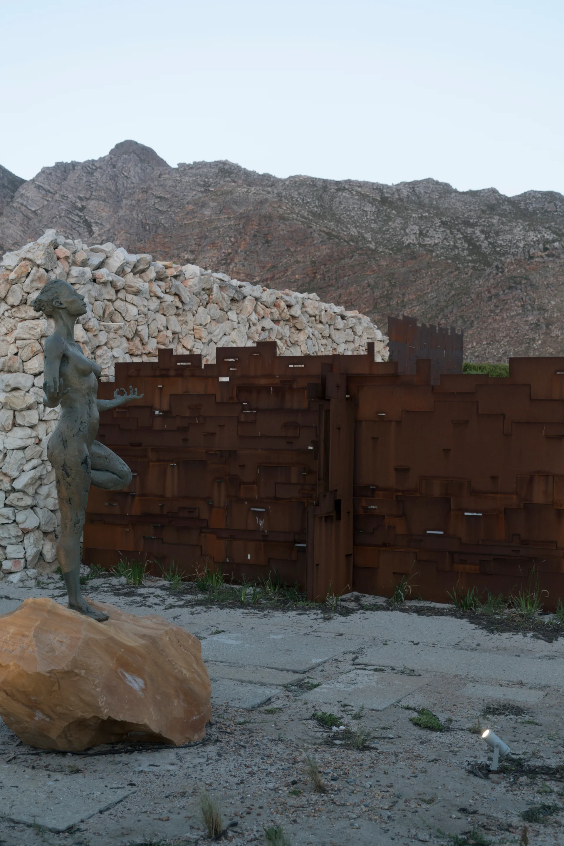 A bronze female sculpture stands on a boulder before a geometric rusted metal screen and stacked stone wall with mountains in the background.
