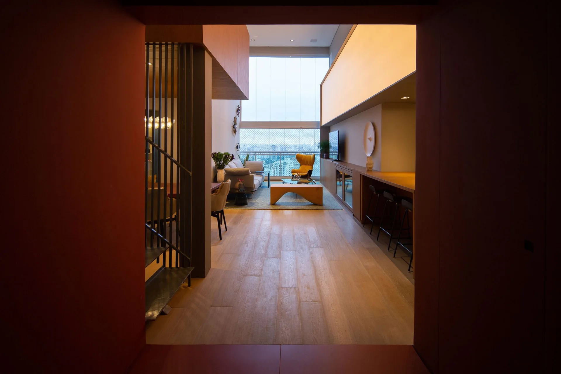 A double-height living room features a grey sofa, yellow wingback chair, curved wood coffee table, and a large illuminated panel over a wooden built-in.