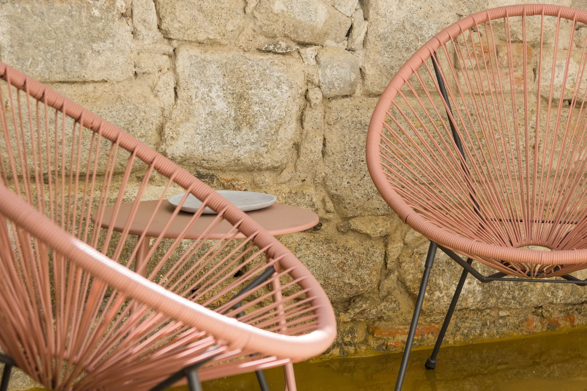 Two dusty pink woven cord lounge chairs and a small matching round metal side table sit in front of a rustic, textured tan stone wall.