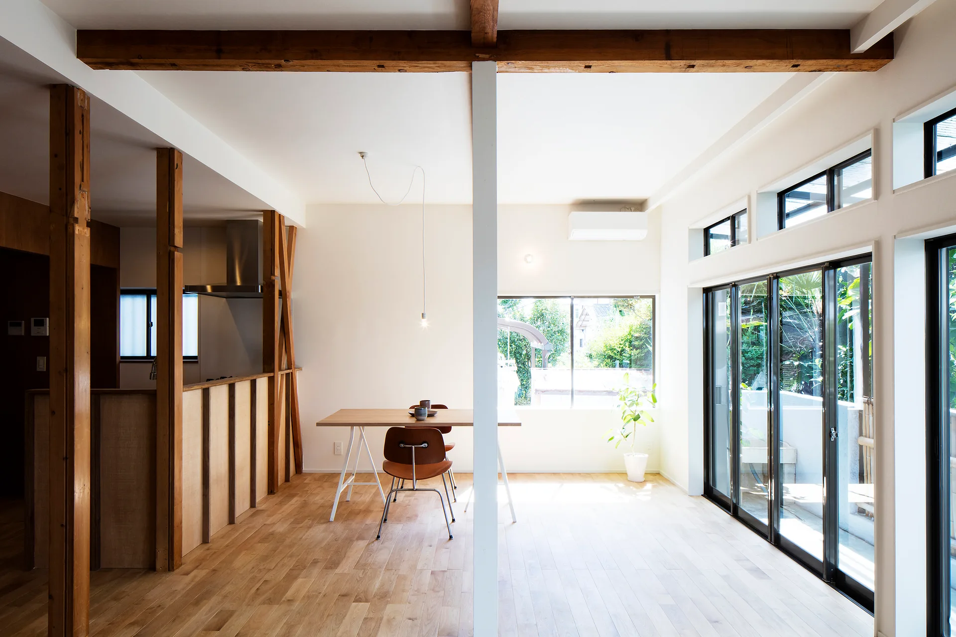 A minimalist dining area featuring a wooden table and chairs under exposed ceiling beams next to large sliding glass doors.