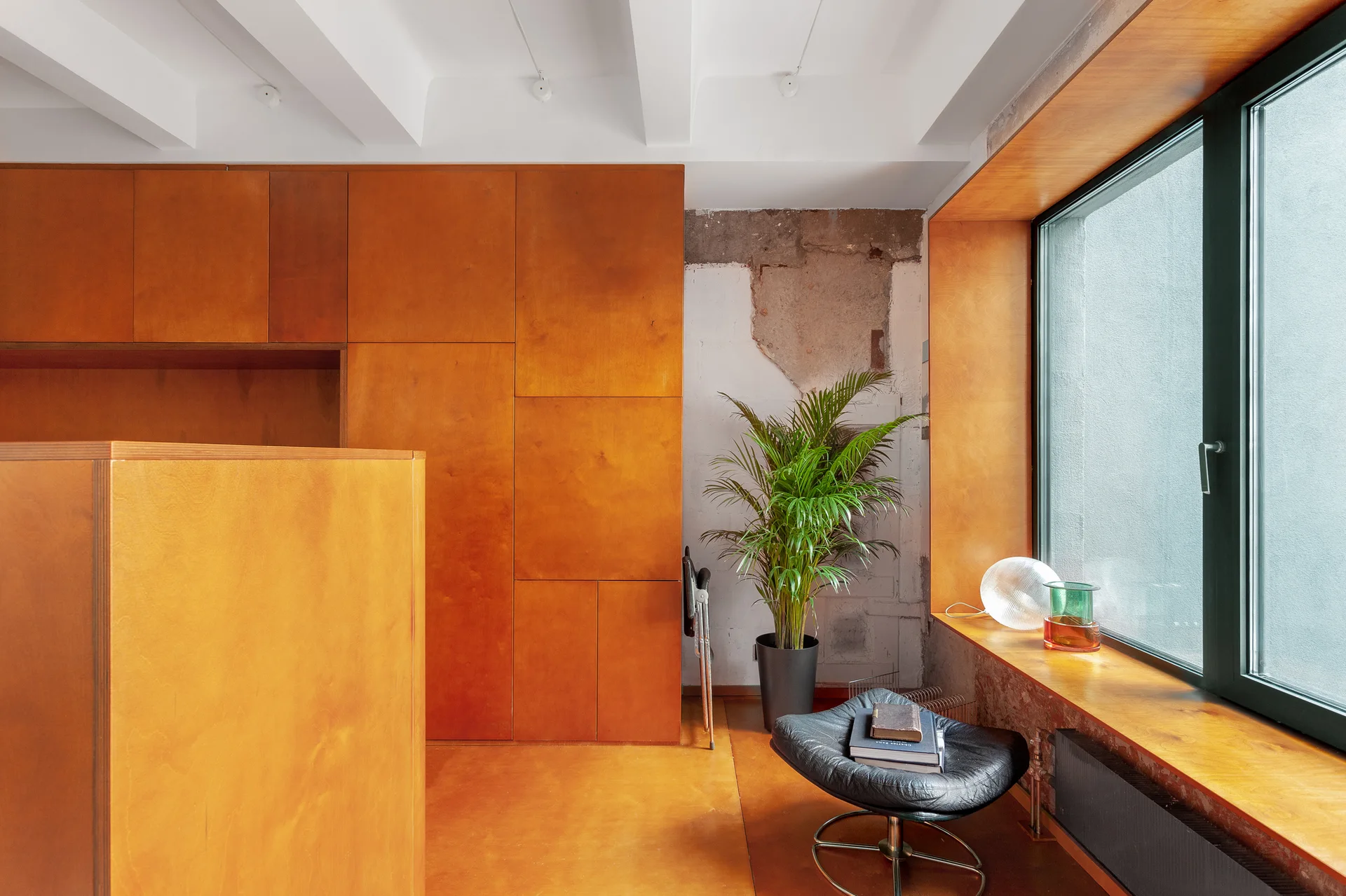 A minimalist interior features warm amber wood wall panels, a wood-wrapped window frame, a black swivel chair, a potted plant, and an unfinished concrete wall.