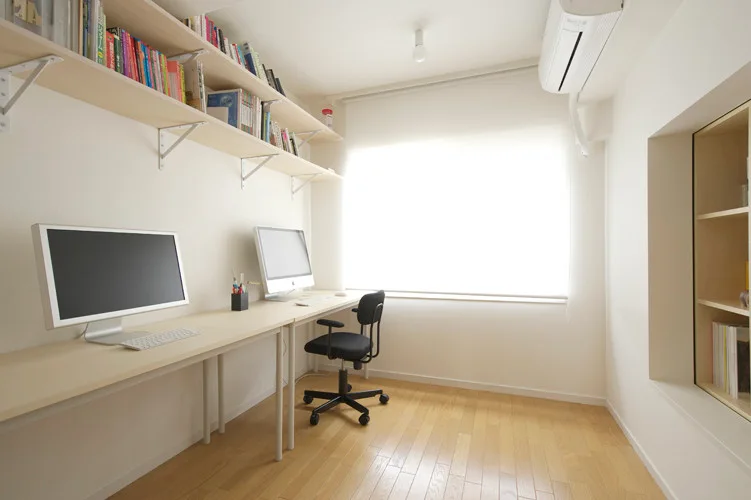 A minimalist home office features a long light wood desk with two computer monitors, overhead floating shelves, and a recessed wall niche.