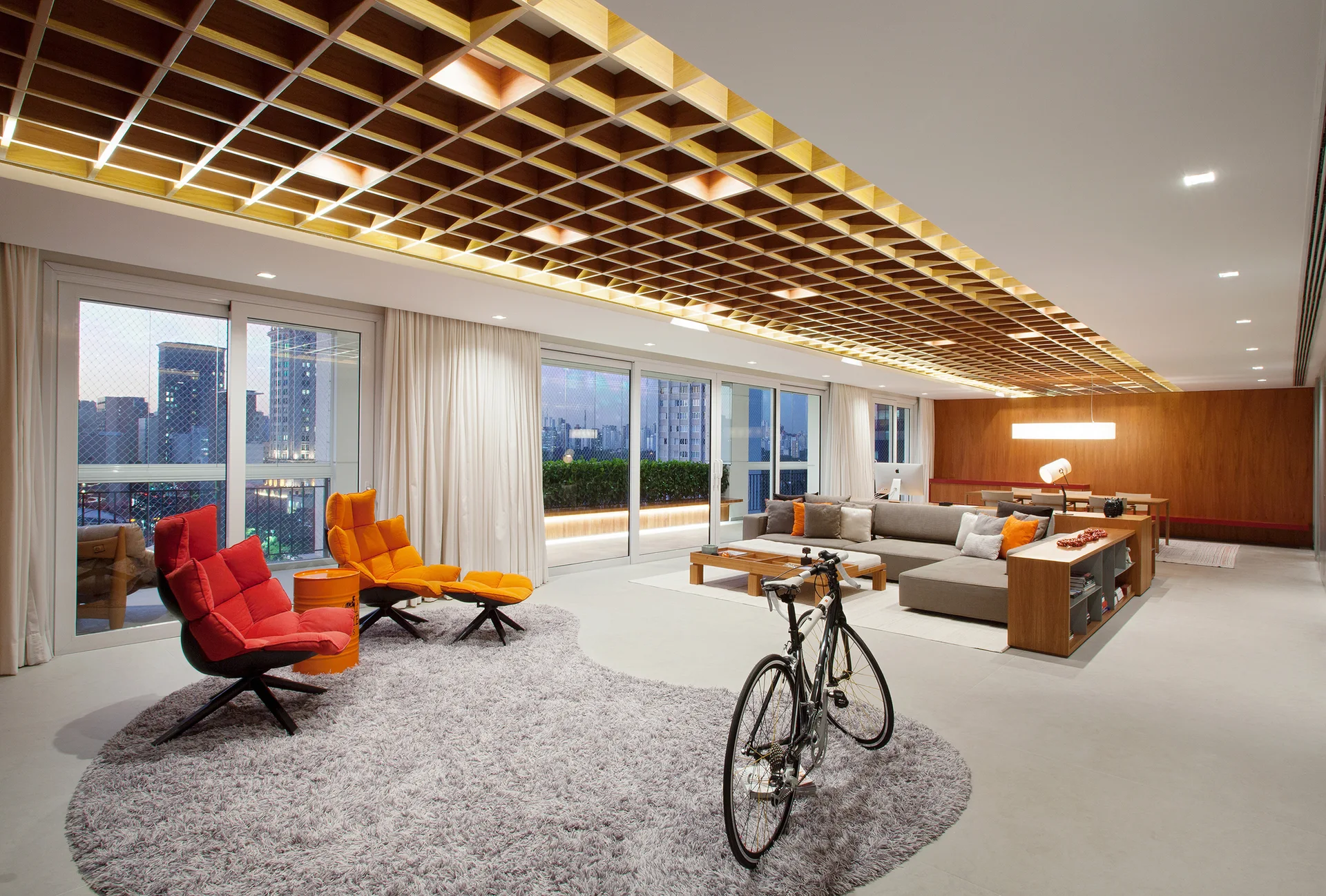 A spacious modern living room featuring a prominent wooden grid ceiling, grey sectional sofa, red and orange accent chairs, and large glass windows.