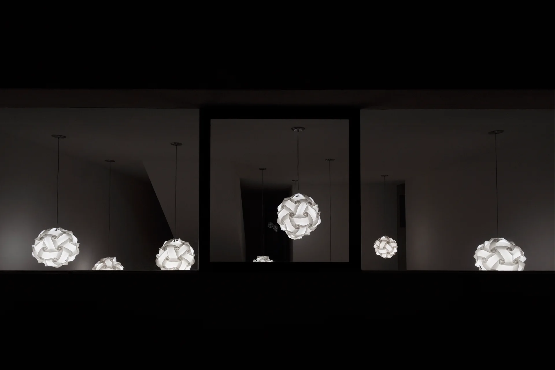 Several glowing white spherical puzzle pendant lights are suspended inside a dark room and reflected in three rectangular window panes.